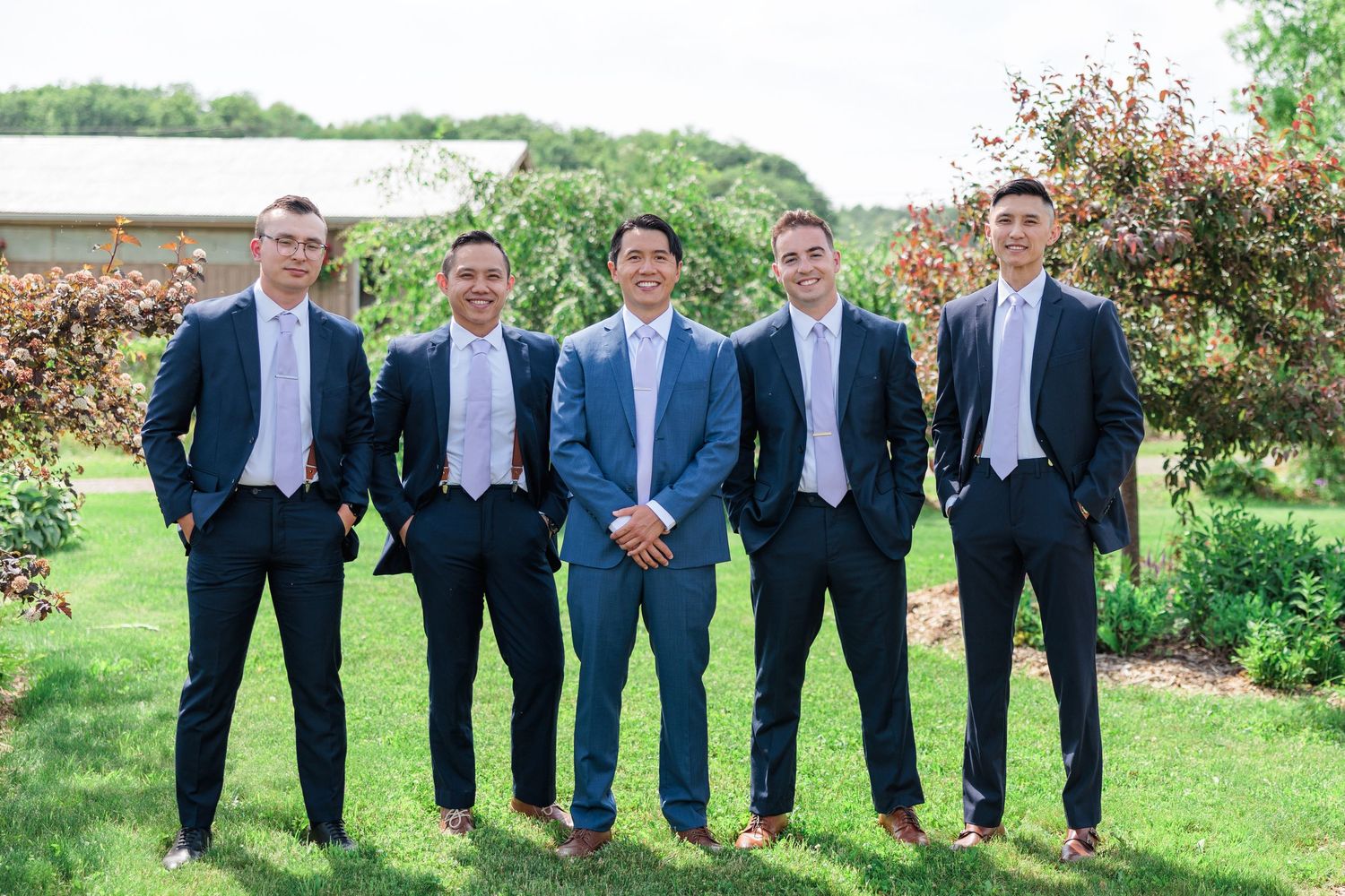 Five groomsmen in navy suits with lavender ties posing outdoors on green lawn.