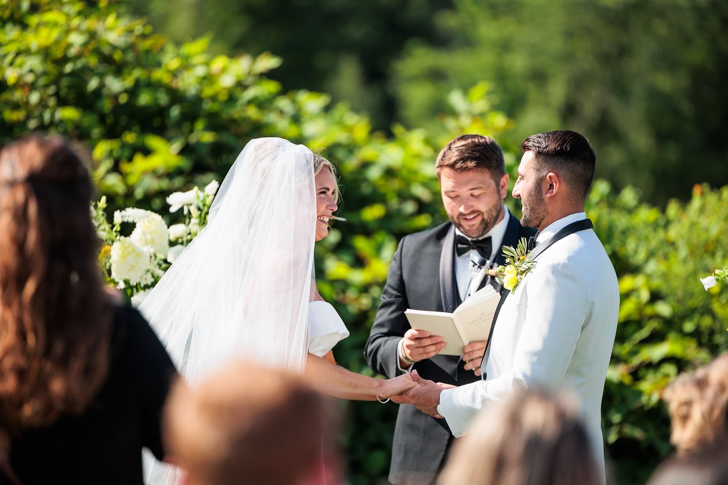 Bride and groom hold hands while officiant reads vows at sunny outdoor garden wedding ceremony.