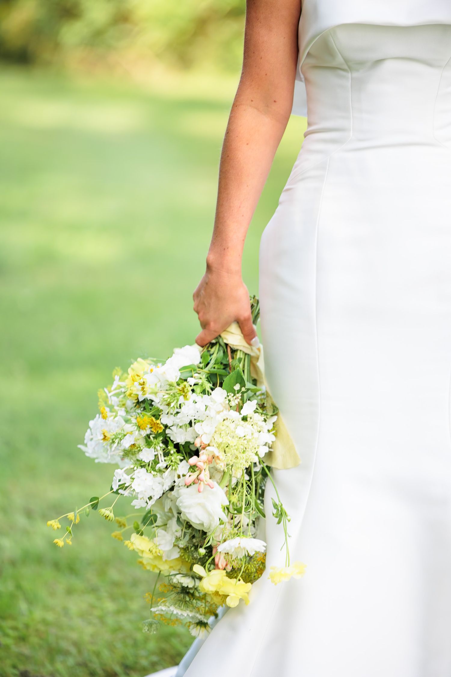Bride in white gown holding a loose bouquet of white and yellow wildflowers outdoors.