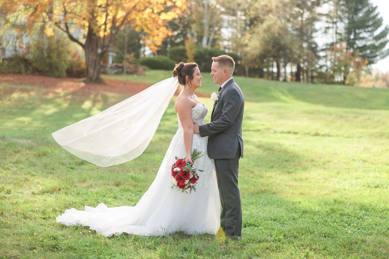 Bride and groom standing in green field with veil flowing and colorful autumn foliage behind.
