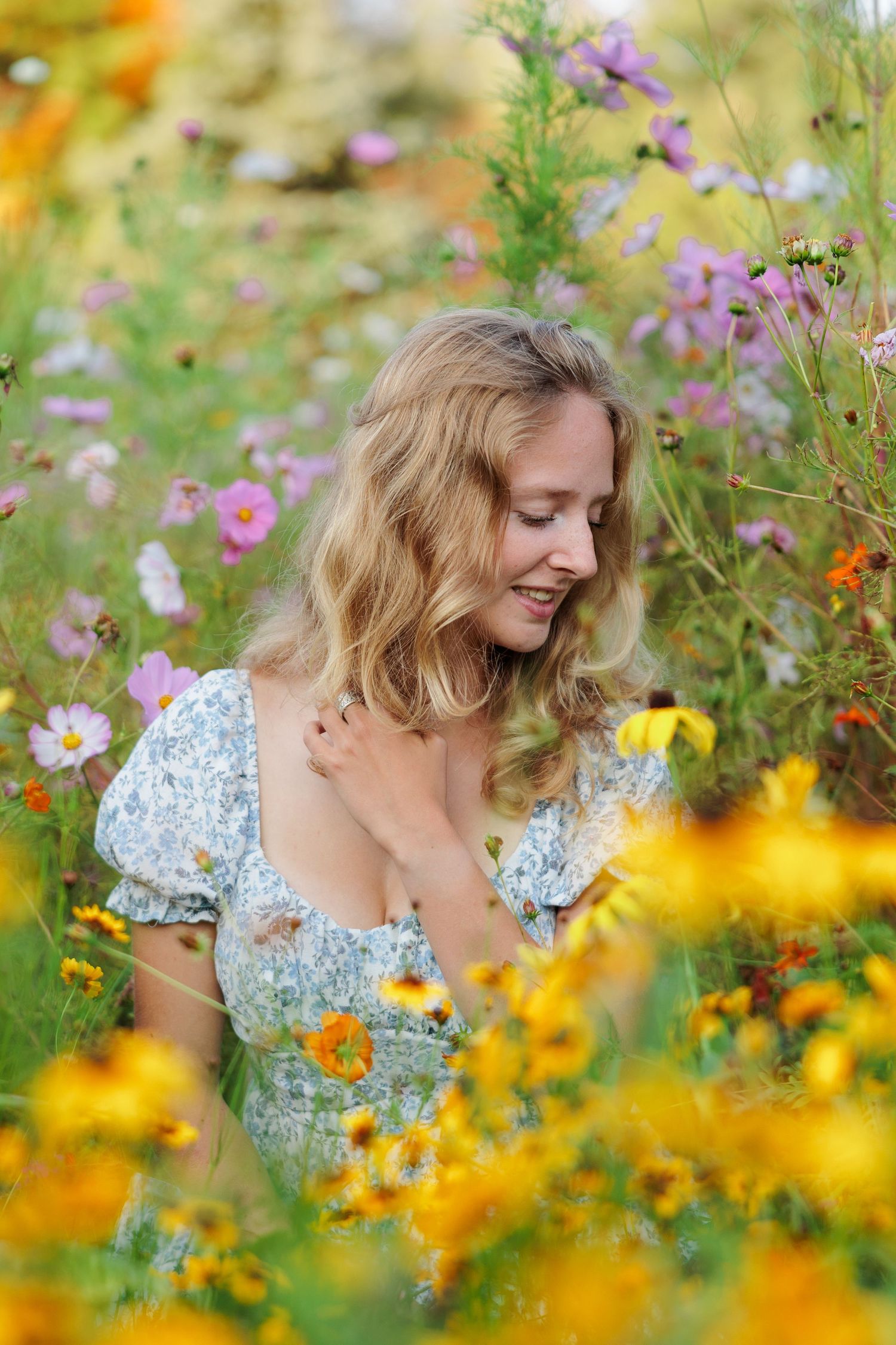 A smiling blonde woman in a floral dress sits among vibrant wildflowers including yellow and pink blooms in a garden.