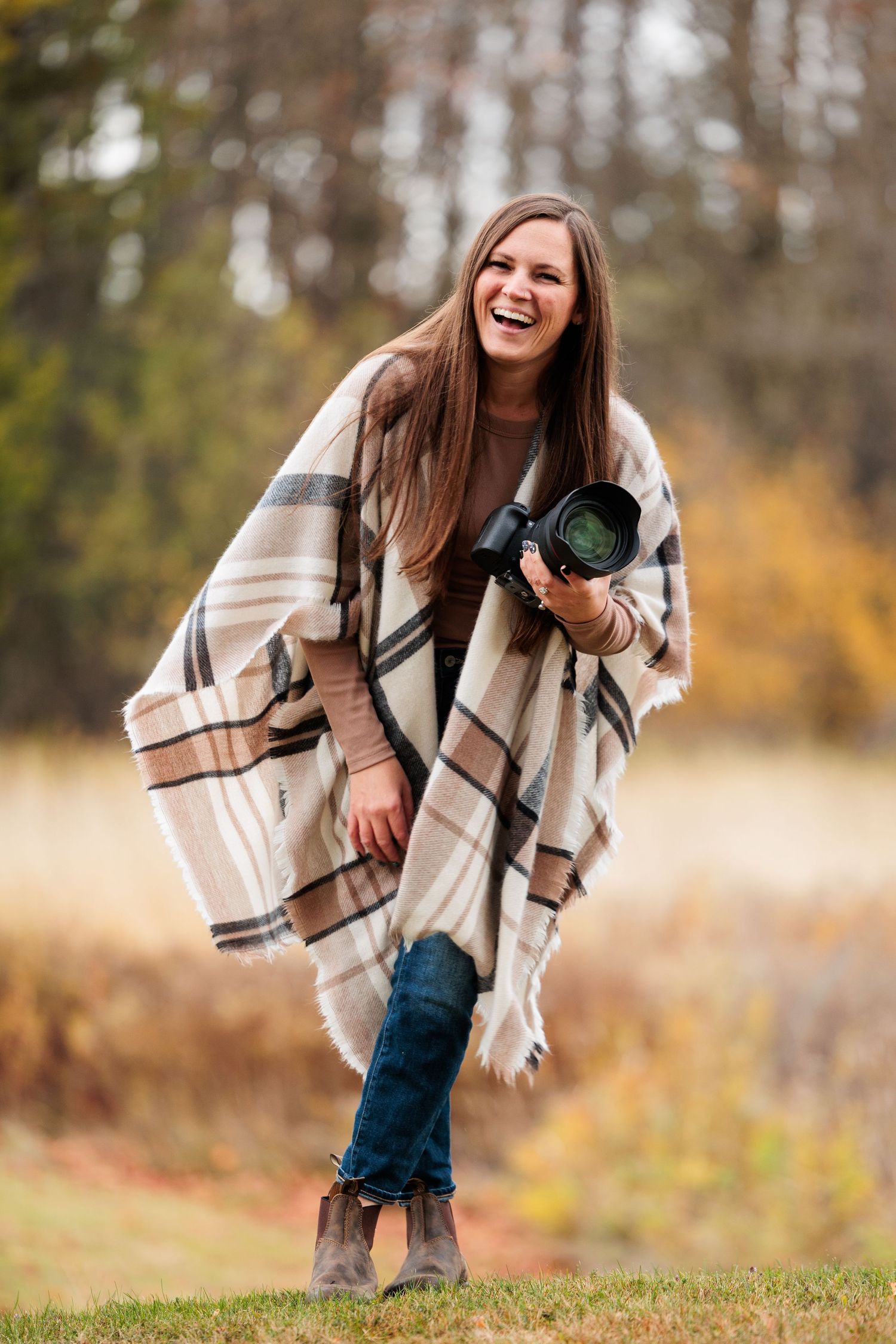 Laughing woman wrapped in plaid blanket scarf holding a camera outdoors in autumn forest setting.
