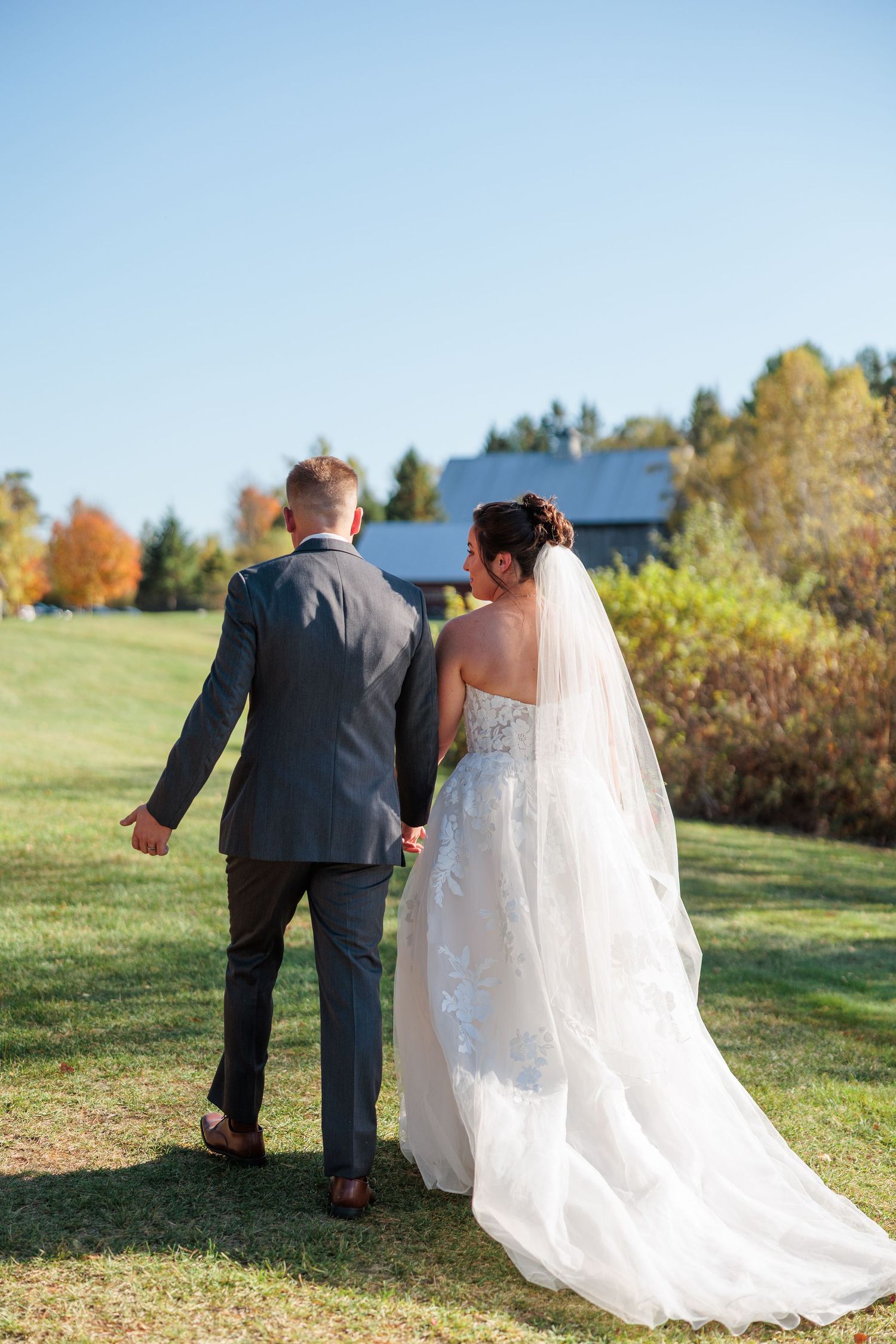 Couple walks across a grassy hilltop on their wedding day with a flowing white dress trailing behind.