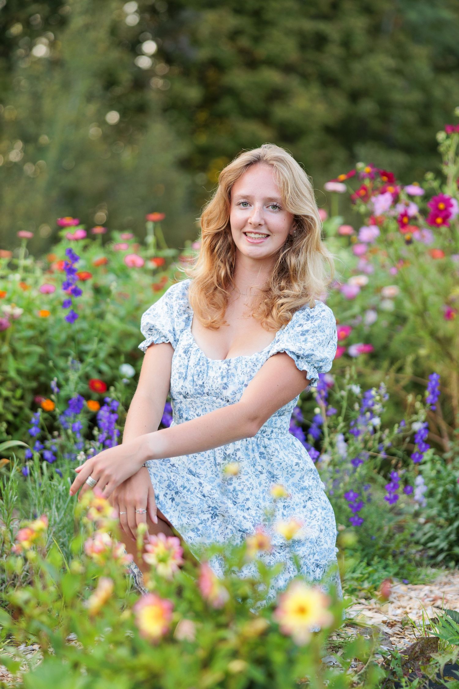 Young blonde woman in floral blue dress smiling in a vibrant garden filled with colorful blooms.
