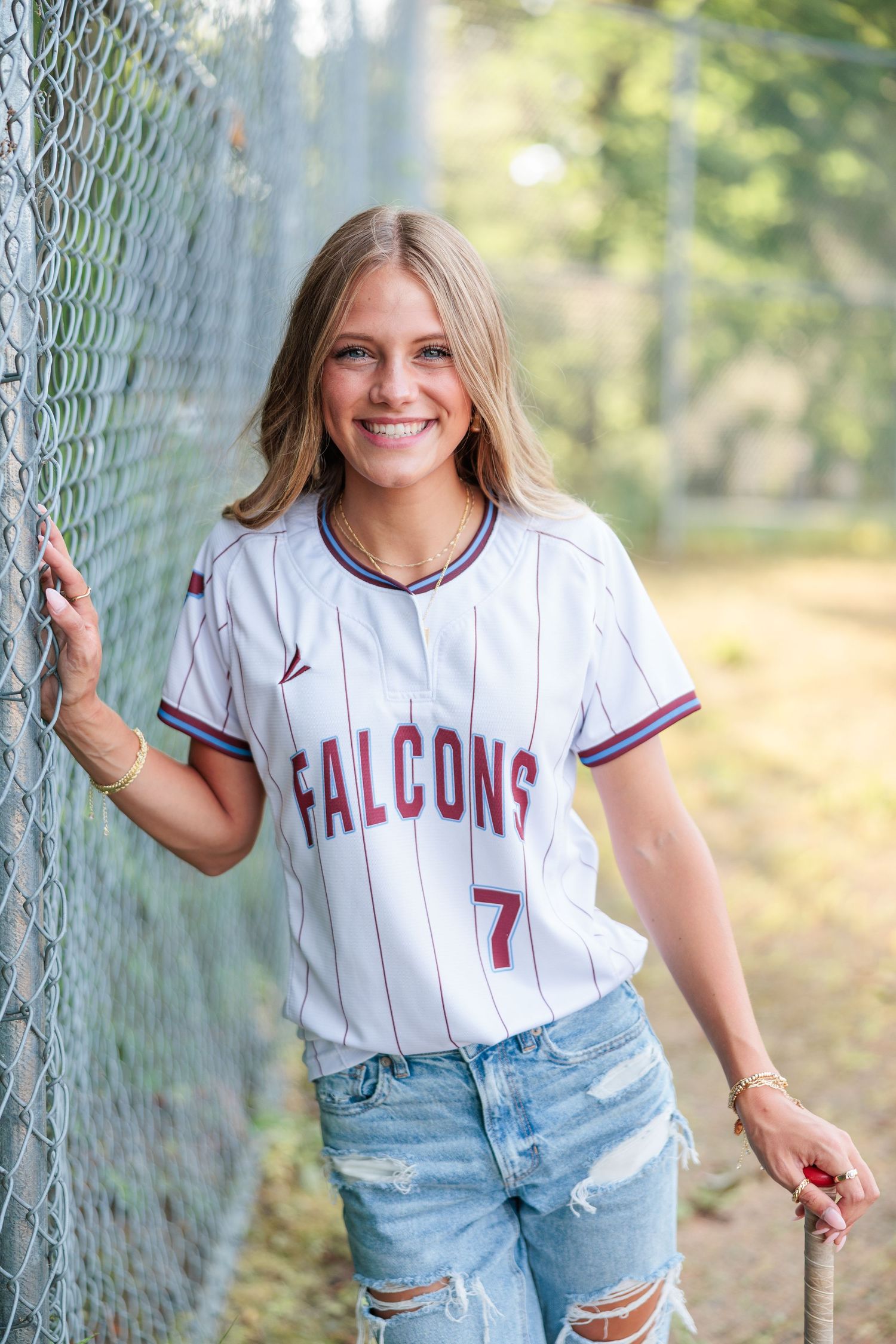 Smiling teen girl in Falcons #7 baseball jersey and ripped jeans leaning against chain-link fence outdoors.