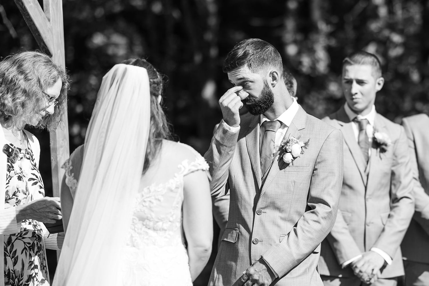 Tearful groom wipes eyes during emotional outdoor wedding ceremony while bride stands before him.