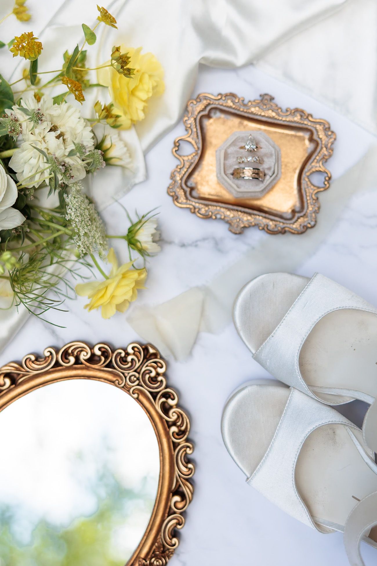Bridal flatlay with yellow flowers, gold mirror, white sandals, and diamond wedding rings on tray.