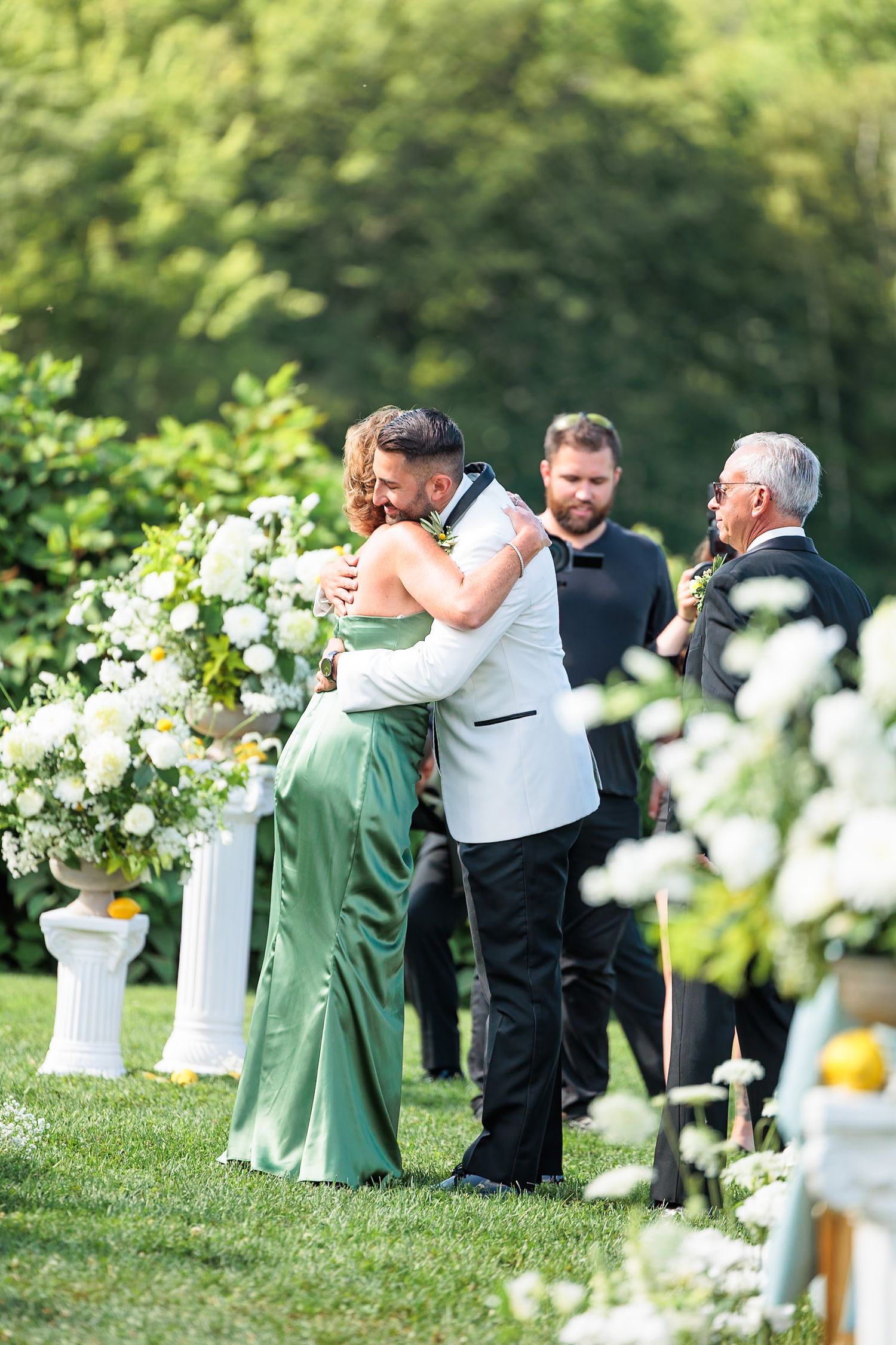 Groom in white tuxedo embracing a woman in green dress at outdoor wedding ceremony.