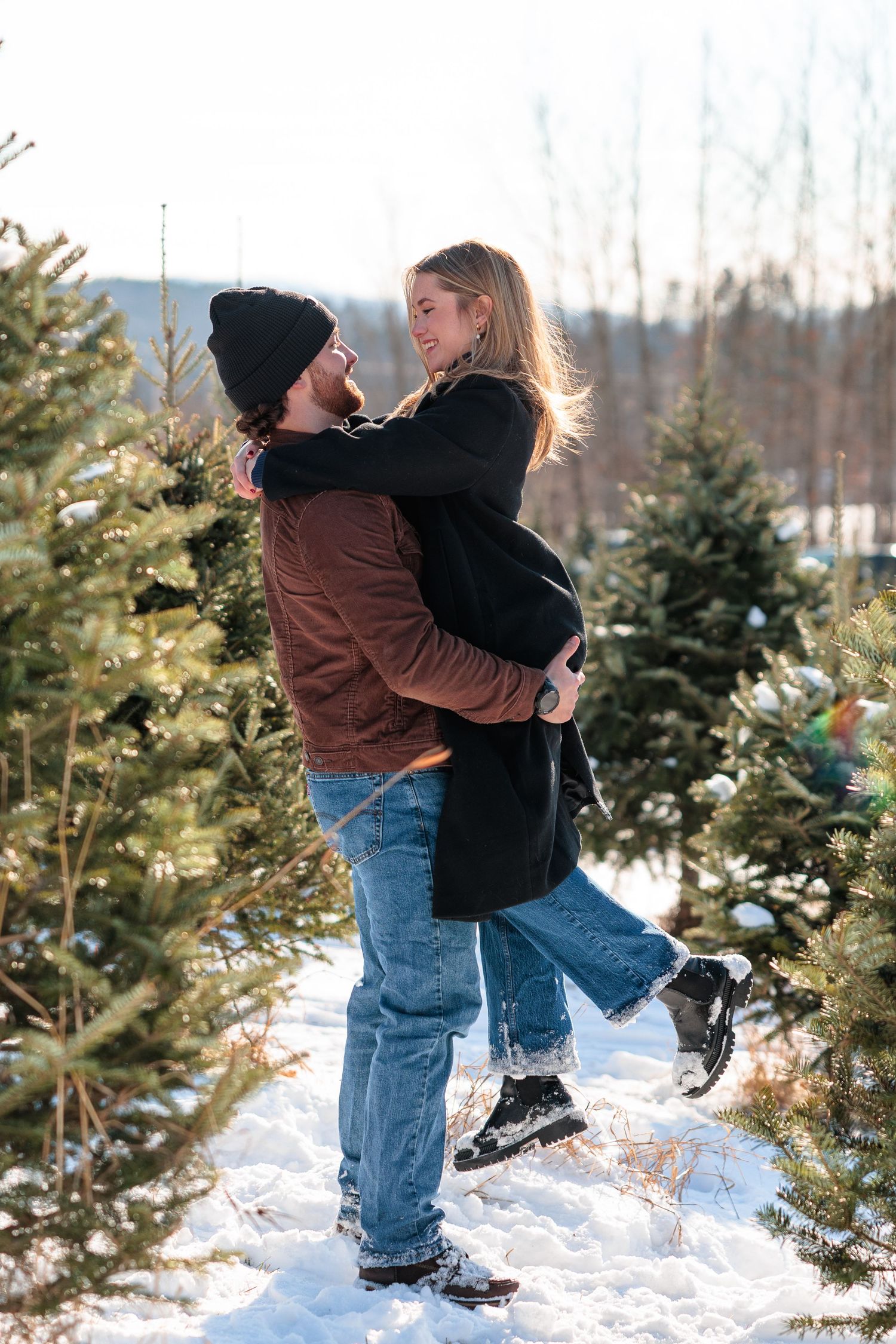 Couple embracing joyfully at a snowy Christmas tree farm, man lifting woman as they smile at each other.
