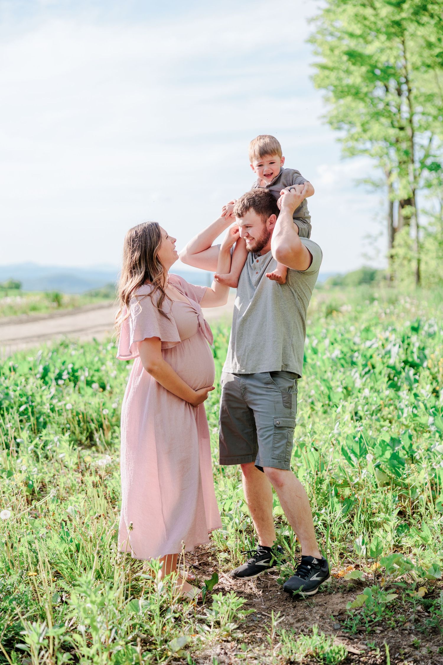 Happy family with pregnant mom, dad, and toddler on shoulders in a field.