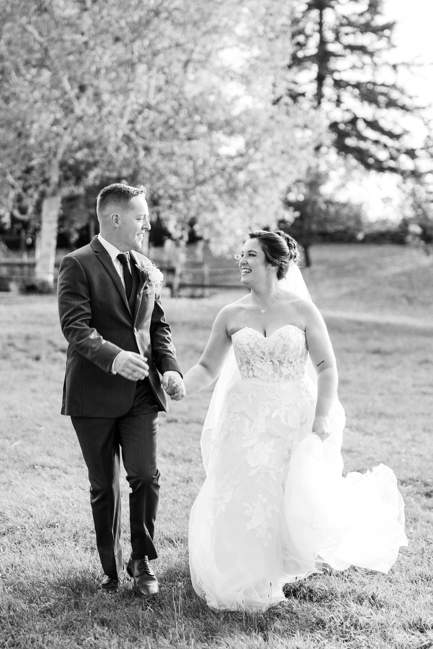 Black and white photo of smiling bride and groom walking together holding hands outdoors.