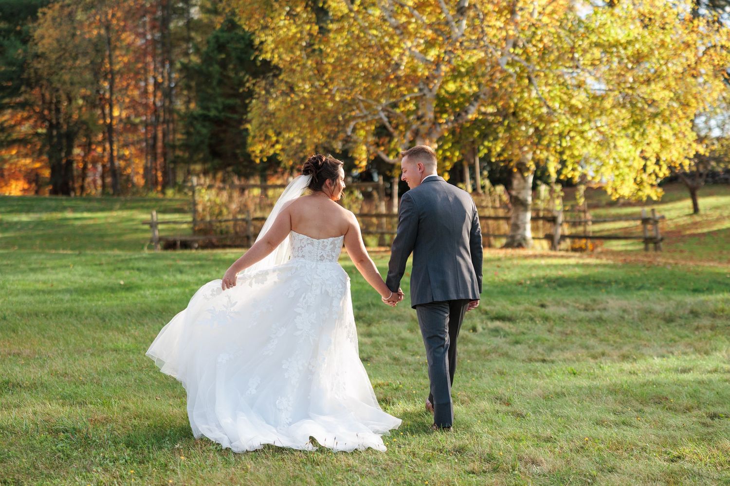 Bride and groom walking hand in hand through a sunlit field with golden autumn trees.