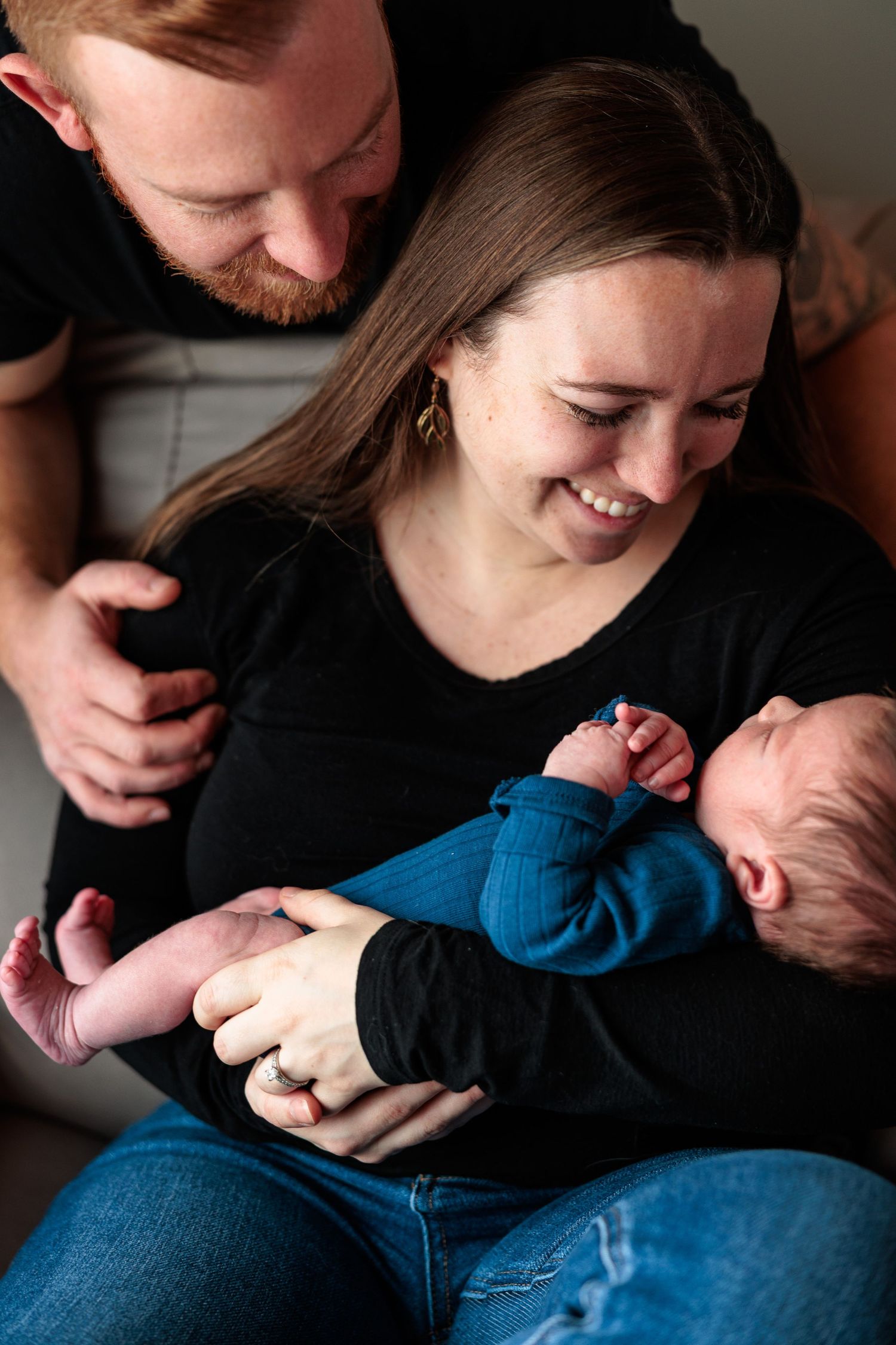 Smiling parents in black shirts lovingly gaze at their newborn baby together.