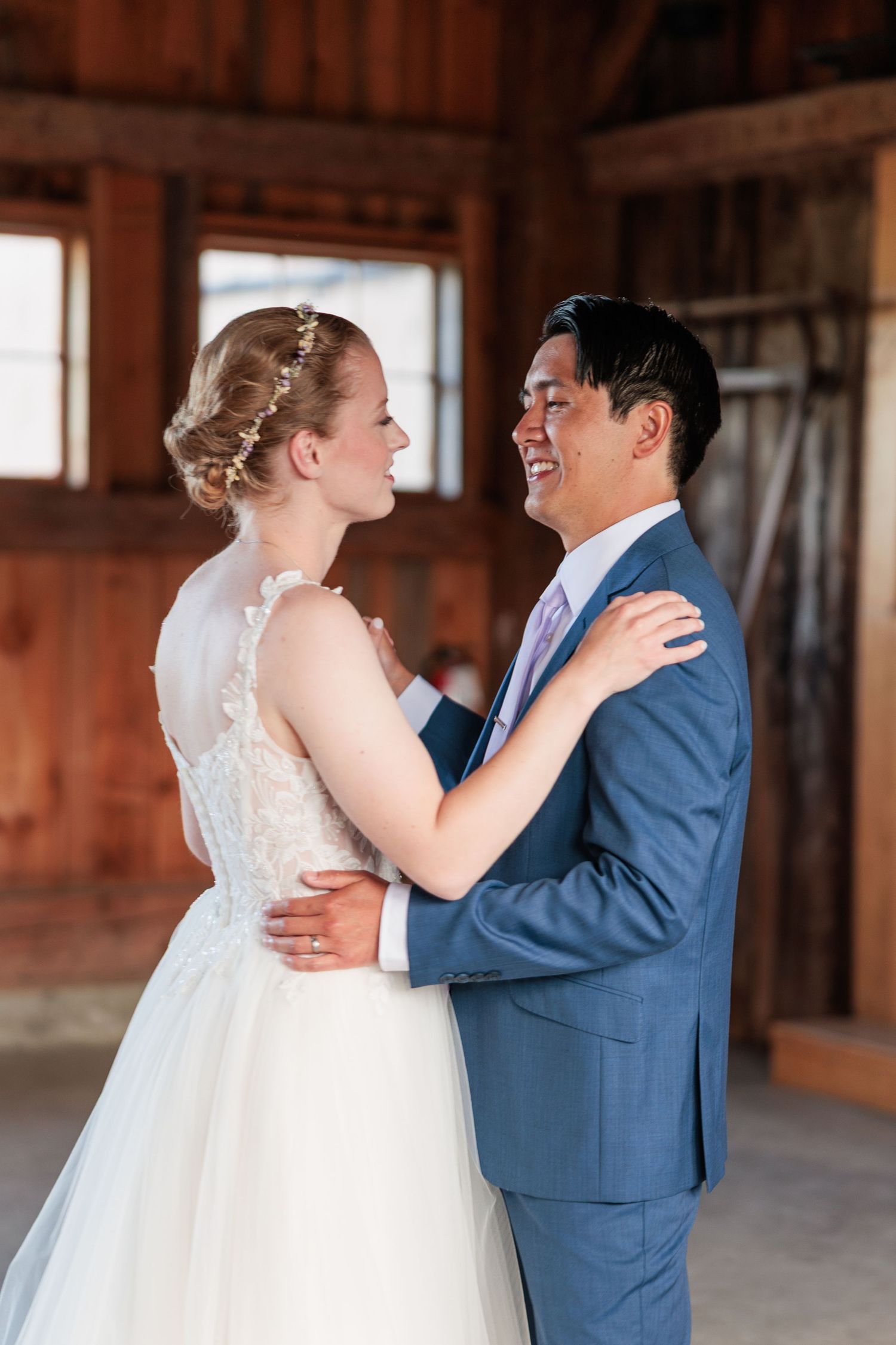 Smiling groom in blue suit embraces bride in lace wedding dress during romantic first dance in barn venue.