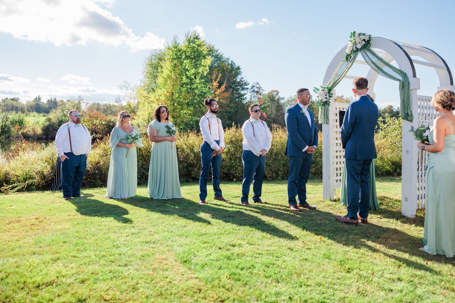 Outdoor wedding ceremony setup with white arch and guests gathered on a sunny hillside.