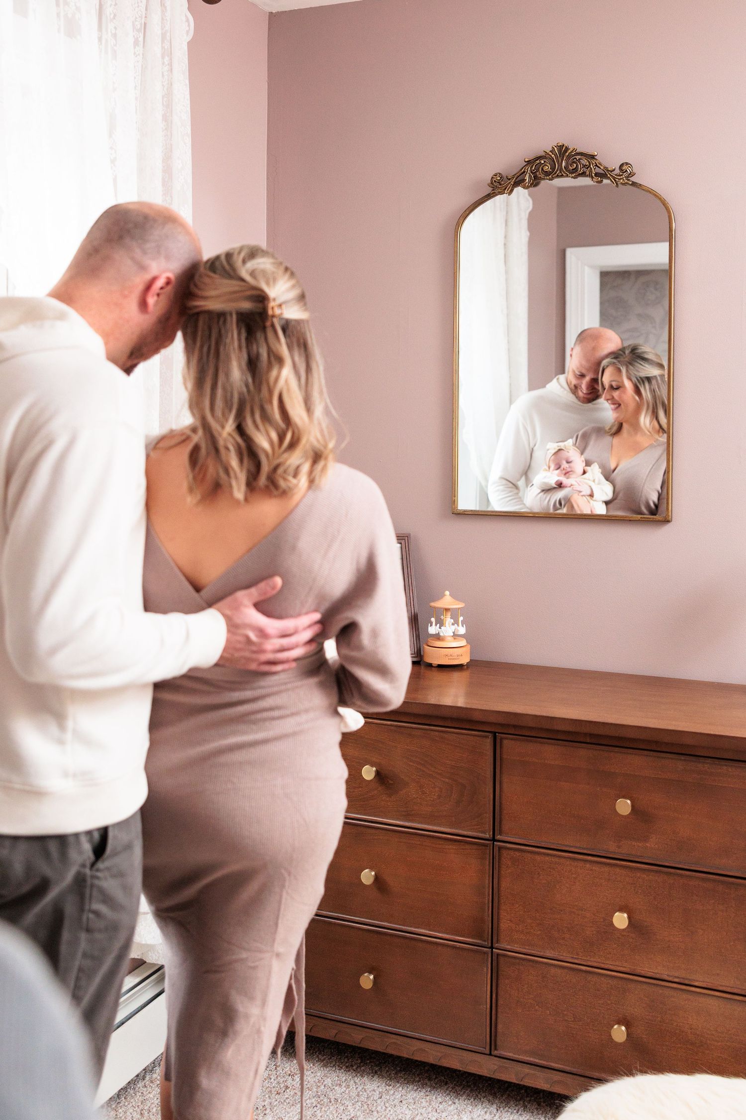 Couple looking at their reflection in ornate mirror mounted above wooden dresser in pink-walled nursery.