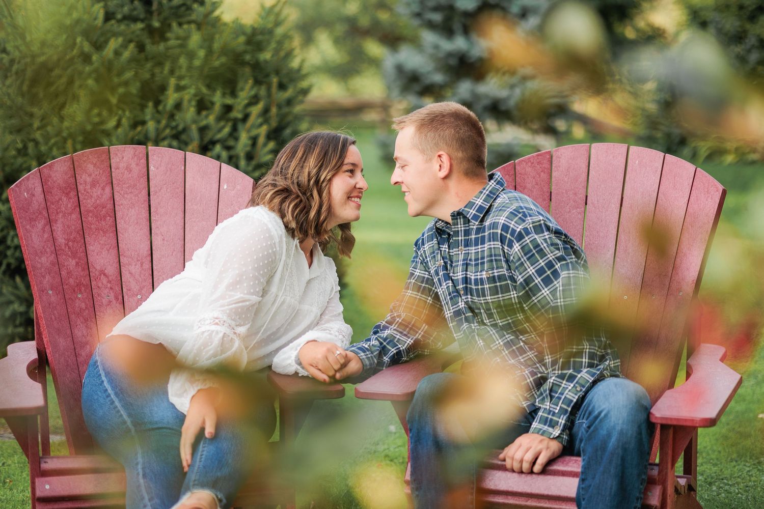 Engaged couple holding hands and smiling at each other while seated in red Adirondack chairs.