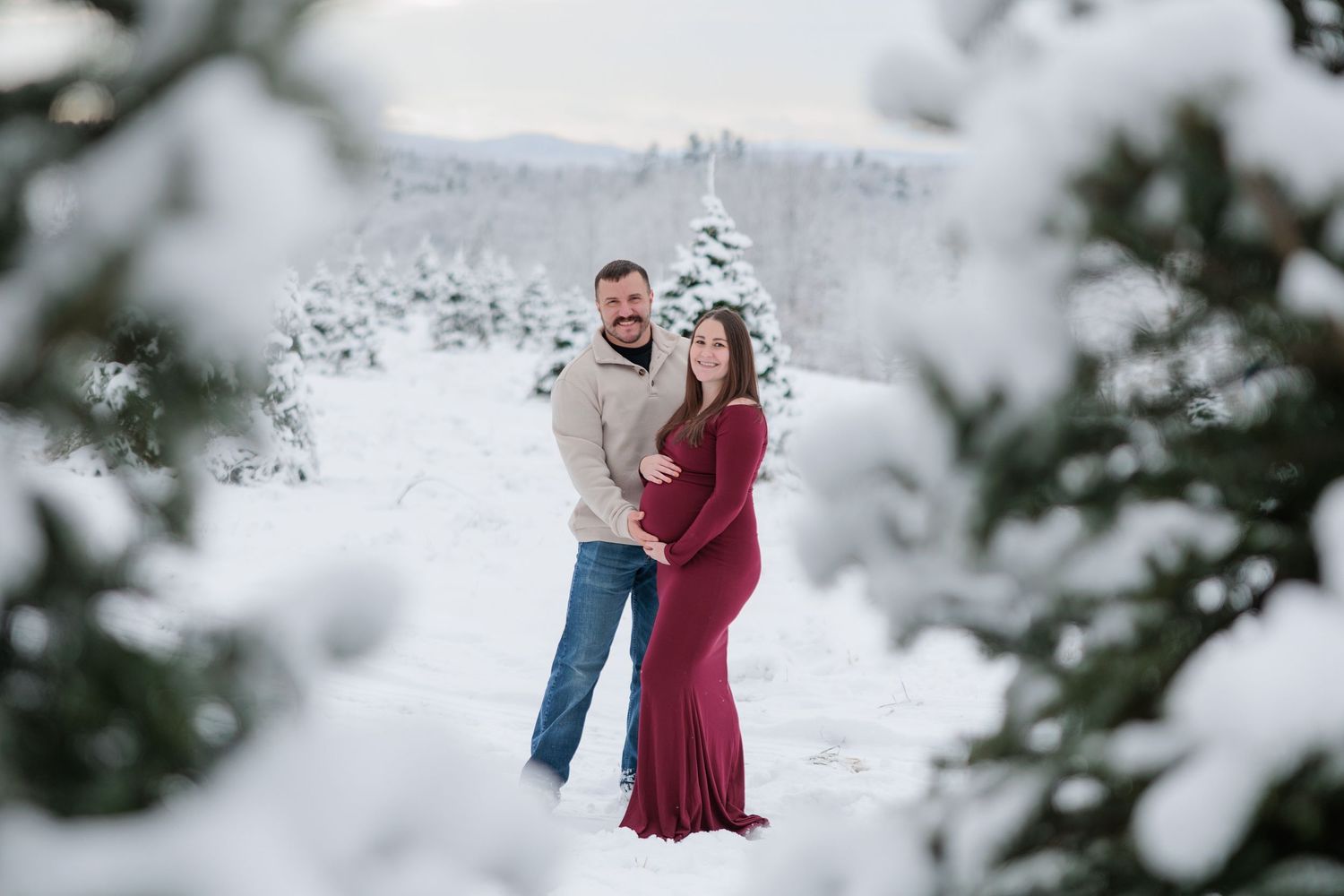 A smiling couple embraces in a winter wonderland surrounded by snow-dusted evergreen trees.