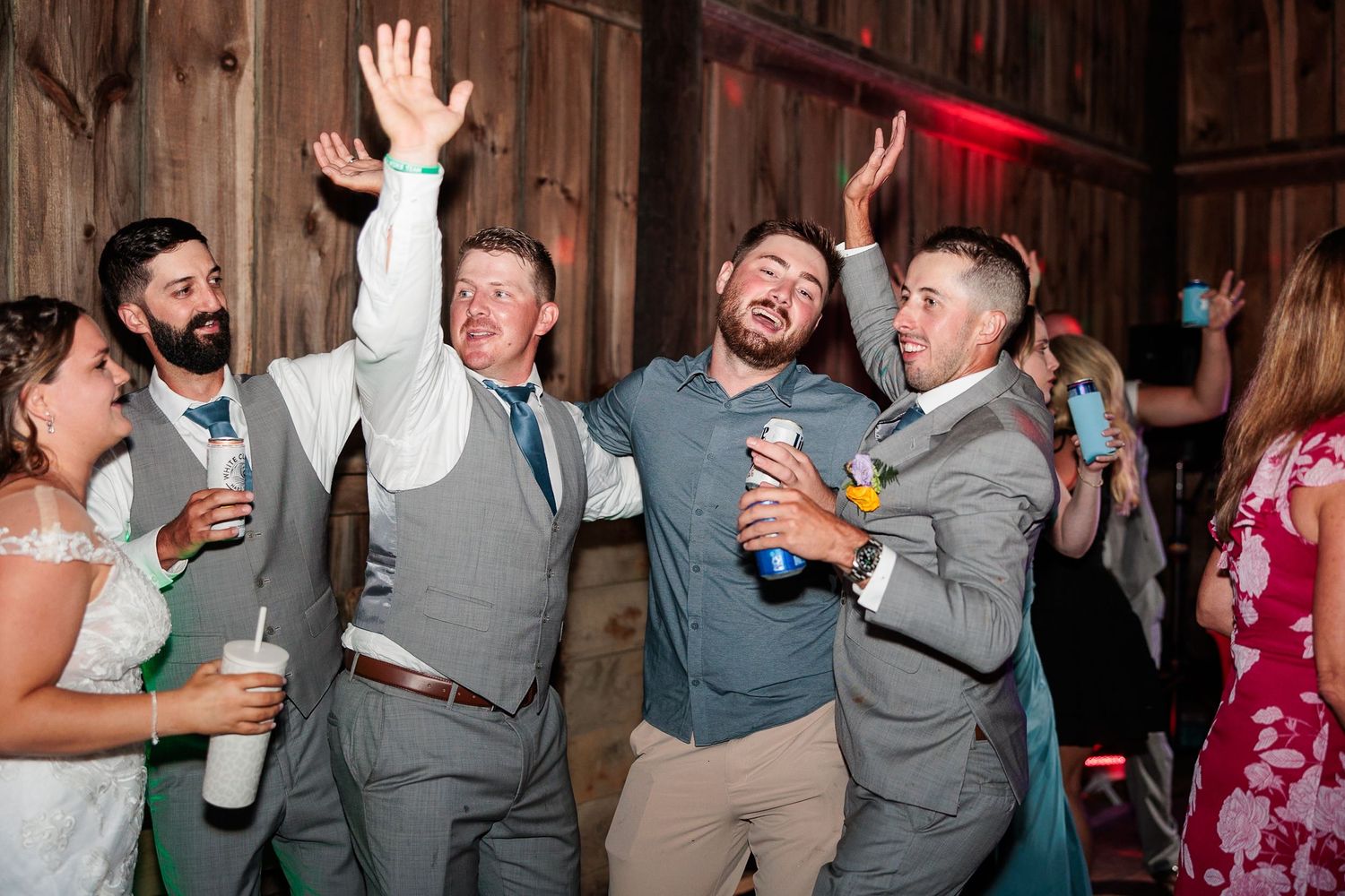 Groom and groomsmen in gray suits cheer and dance together joyfully at wedding reception.