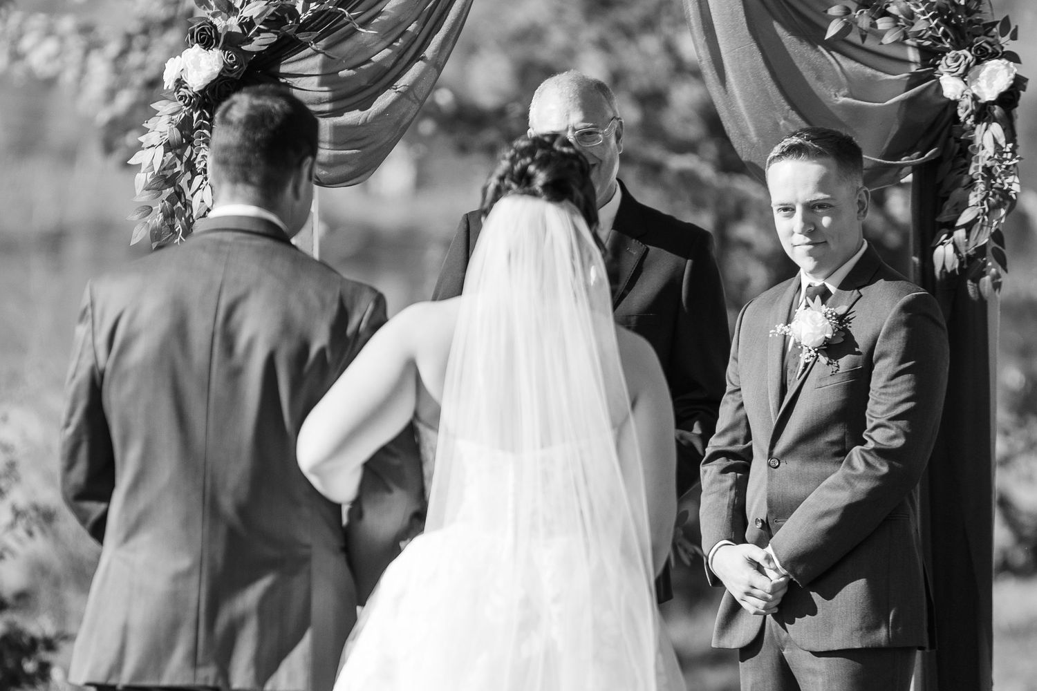 Black and white photo of bride and groom exchanging vows at floral arch during outdoor wedding ceremony.