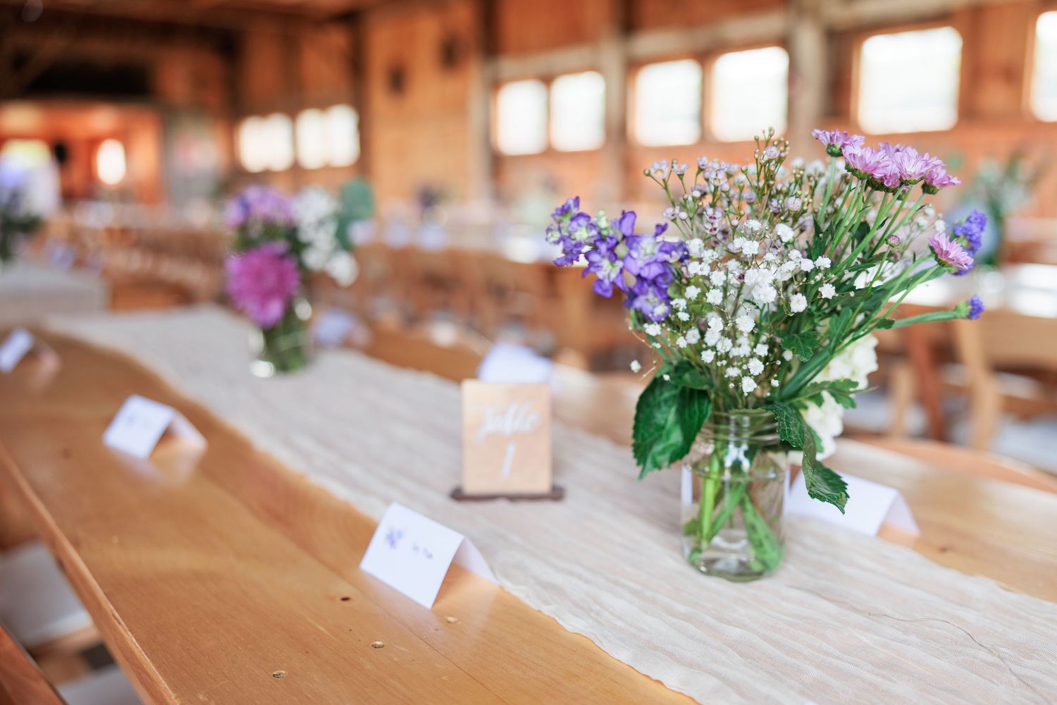 Mason jar centerpieces with purple wildflowers and baby's breath on rustic wooden tables in a barn wedding venue.