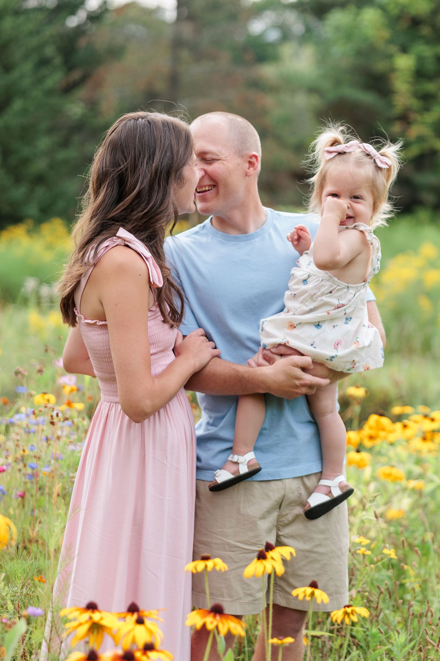 Smiling family with bald father holding toddler daughter in wildflower meadow with yellow blooms.