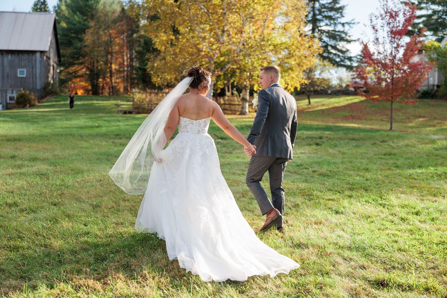 A couple walks through an autumn field with colorful fall foliage and trees in the background.