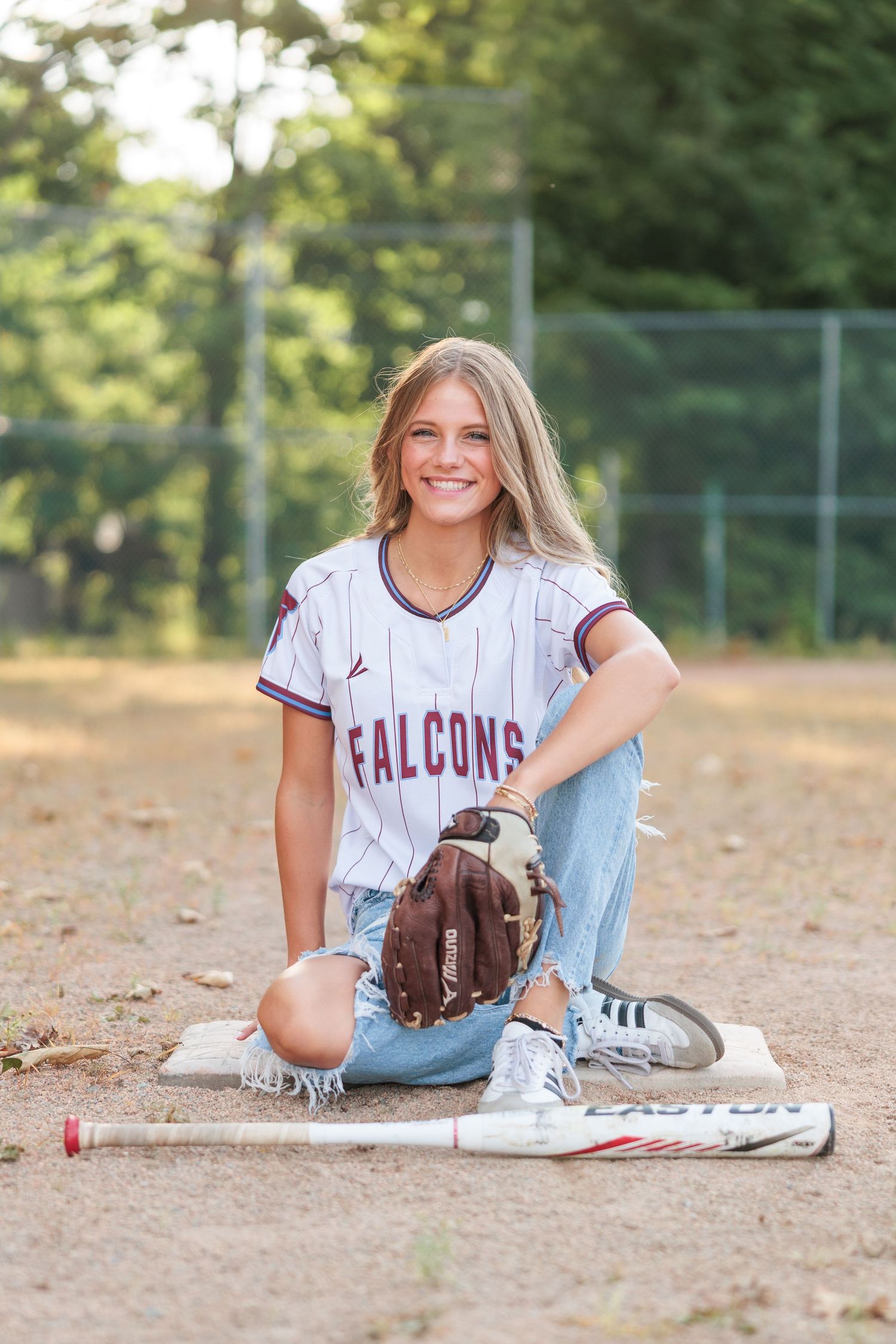 Young woman in Falcons softball jersey poses with glove and Easton bat on baseball diamond.