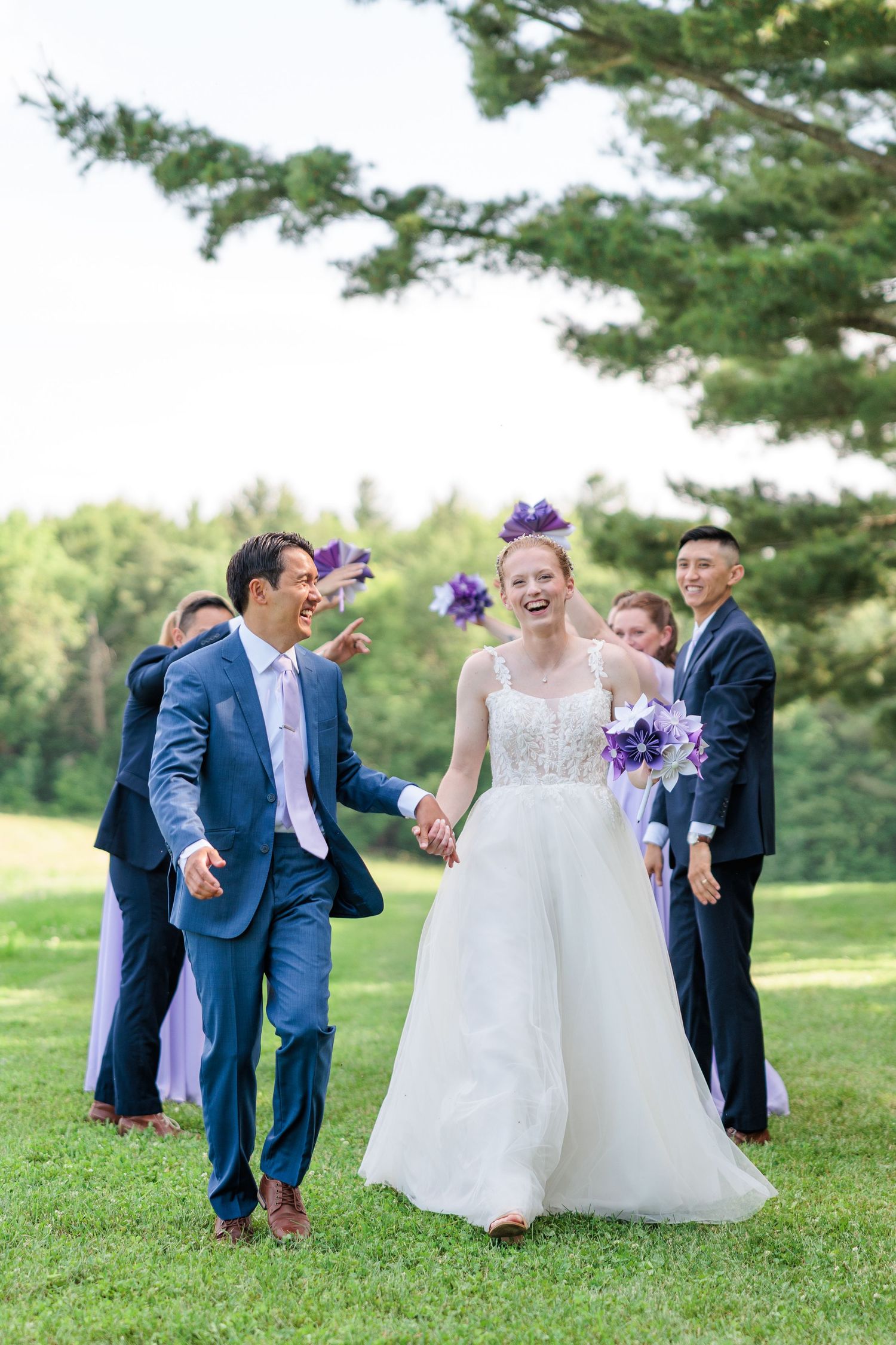 Bride in white lace gown and groom in navy suit walking joyfully with wedding party cheering behind them.