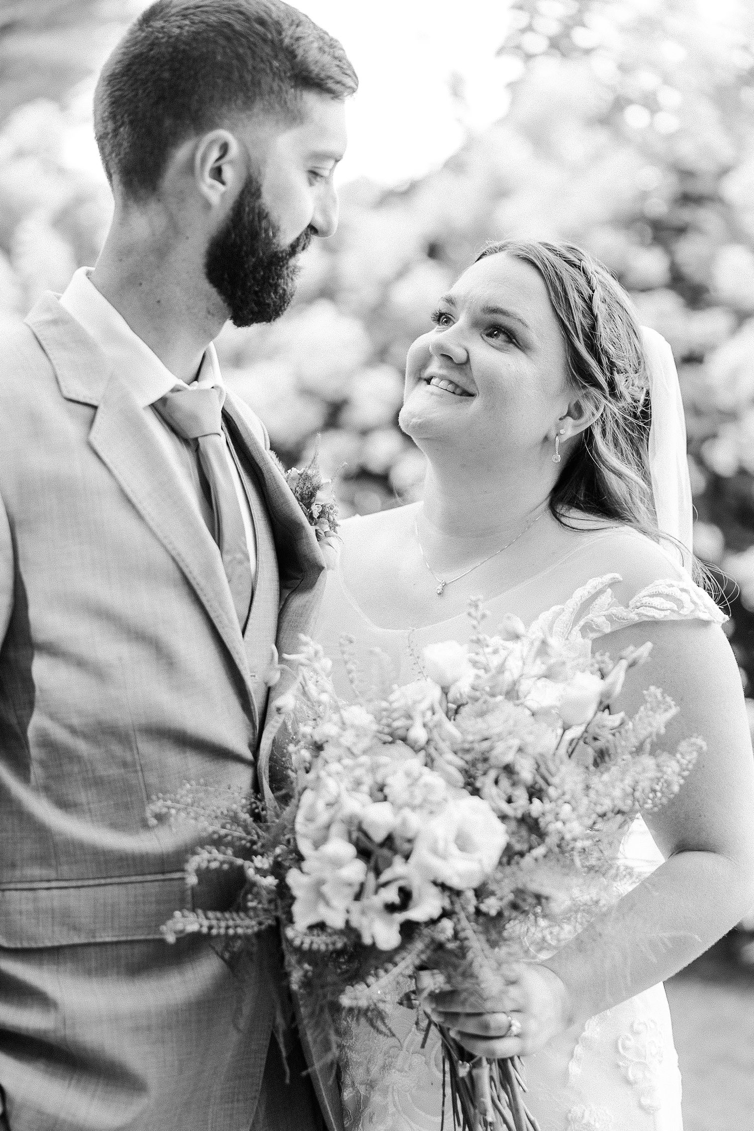 Black and white portrait of smiling bride holding bouquet gazing up at bearded groom in a gray suit.