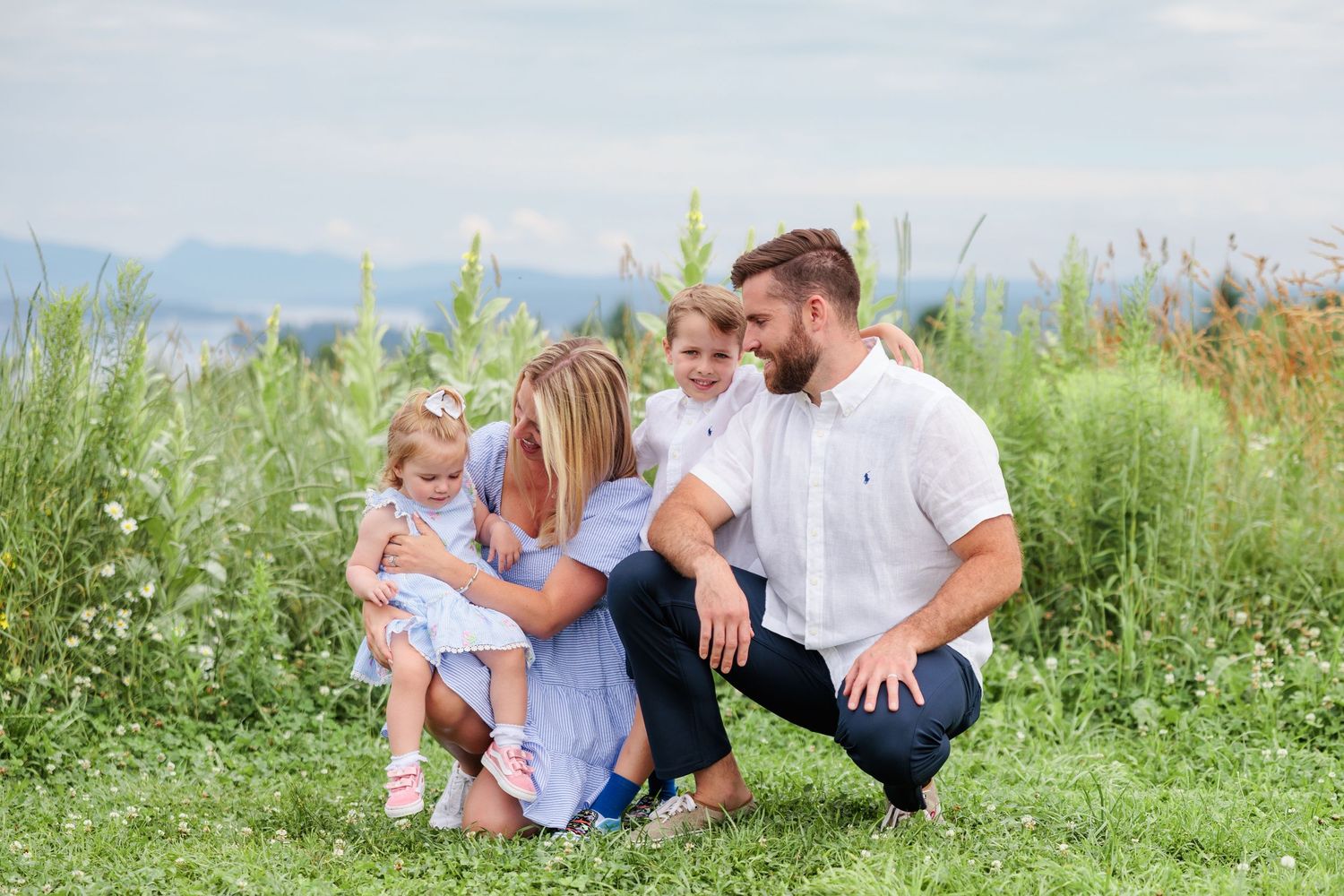 Happy young family of four crouching together in a wildflower field with mountain views.