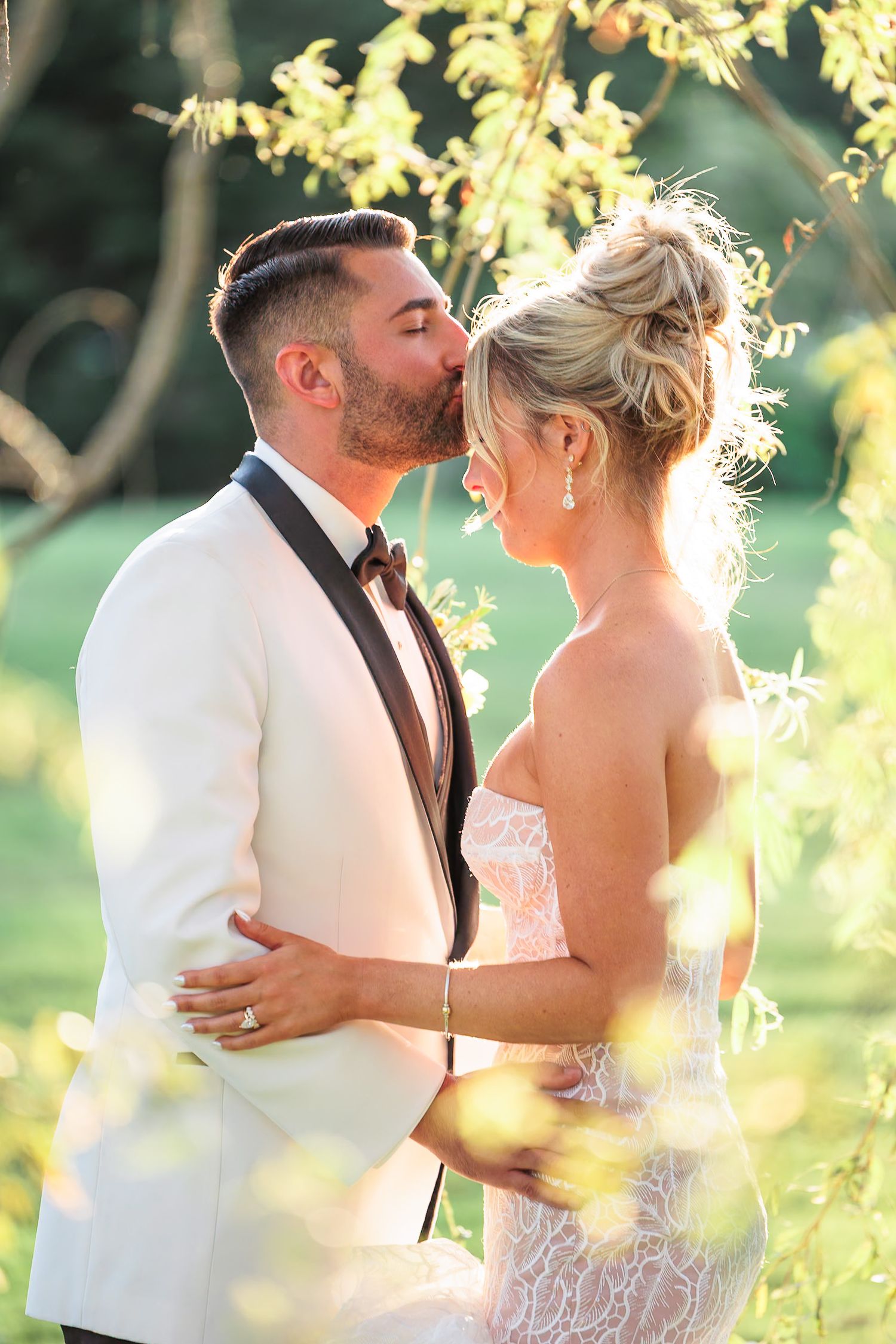 Groom in white tuxedo jacket tenderly kissing bride on cheek during romantic outdoor wedding photo session.