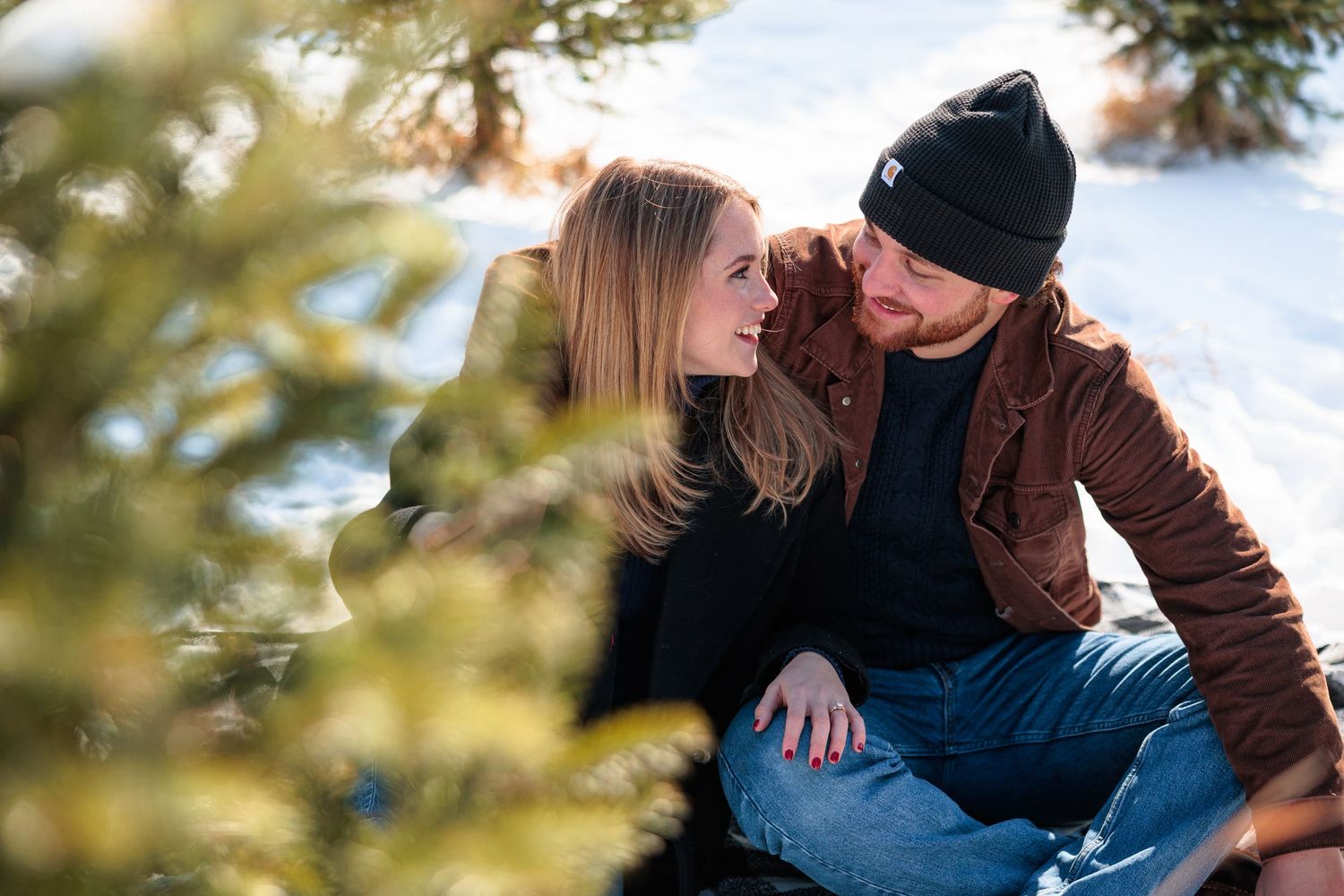 Couple sitting together in the snow at a Christmas tree farm, gazing lovingly at each other.