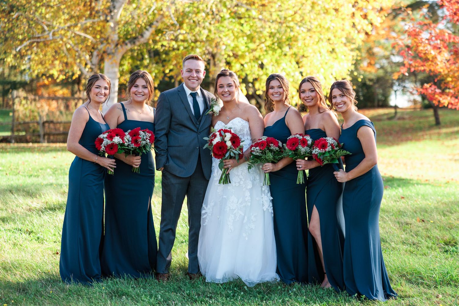 Wedding party members in navy dresses and suits posing on grass with autumn trees in background.