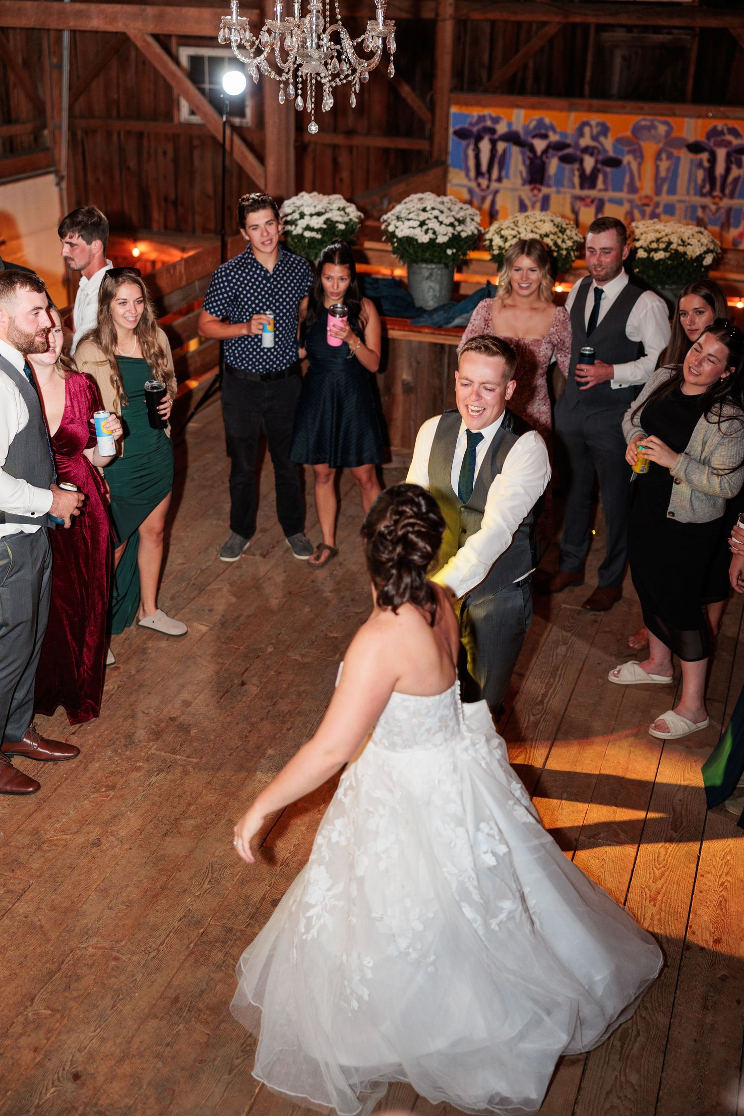 Wedding guests surround newlyweds dancing on wooden barn floor during evening reception celebration.