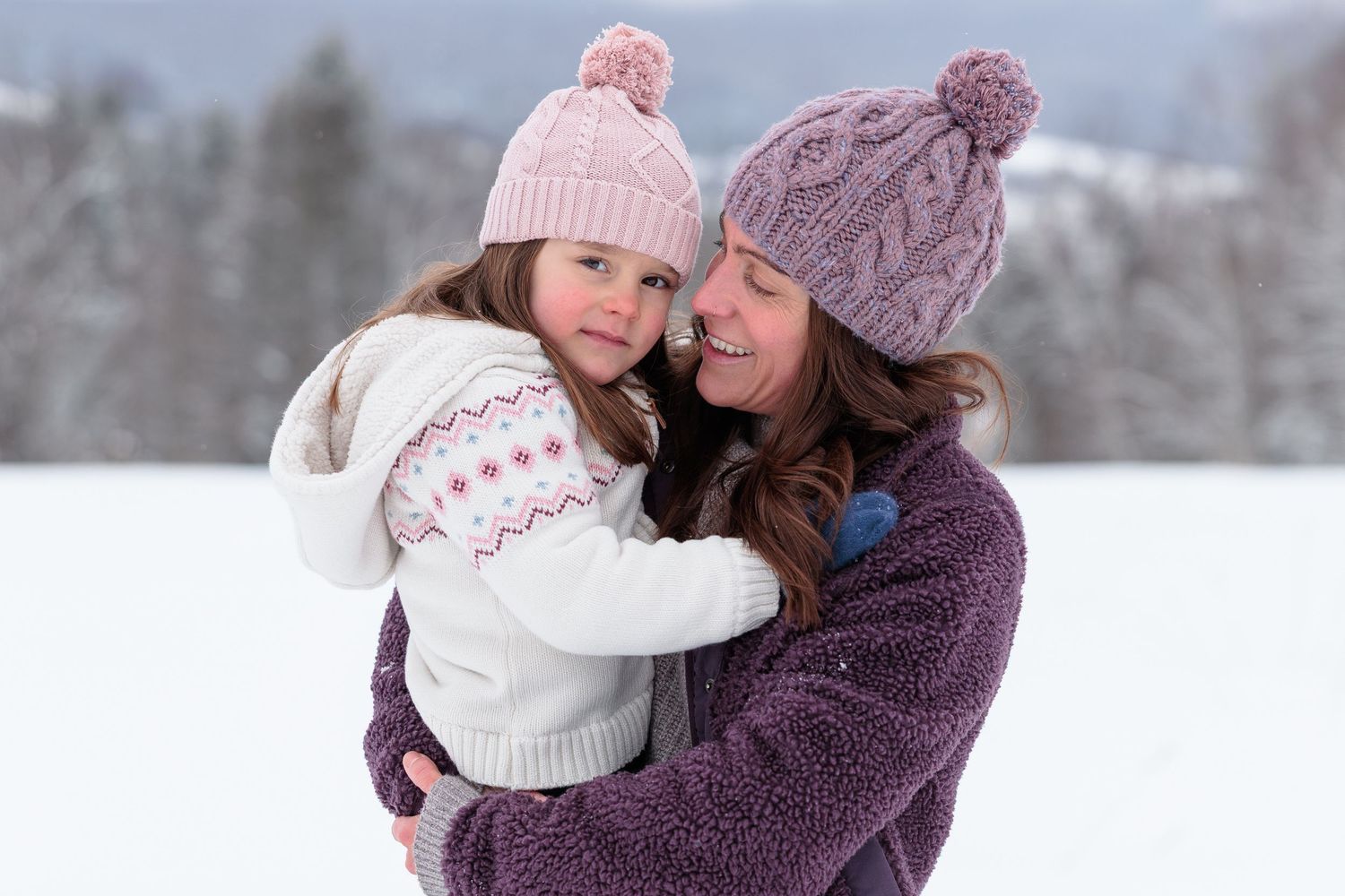 A mother in a knit hat lovingly embraces her young daughter in a snowy winter landscape.