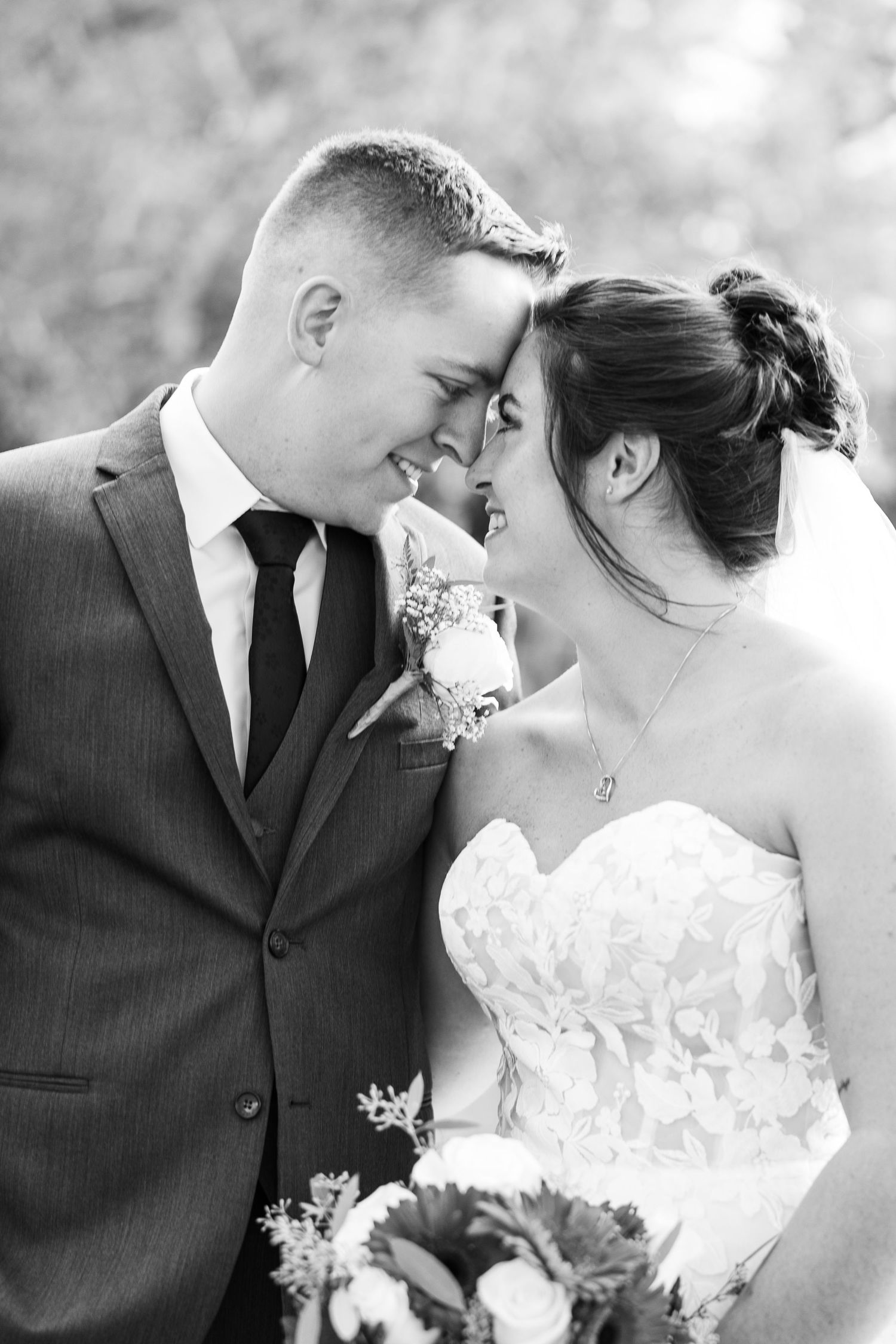 Black and white close-up of bride and groom touching foreheads and smiling tenderly at outdoor wedding.
