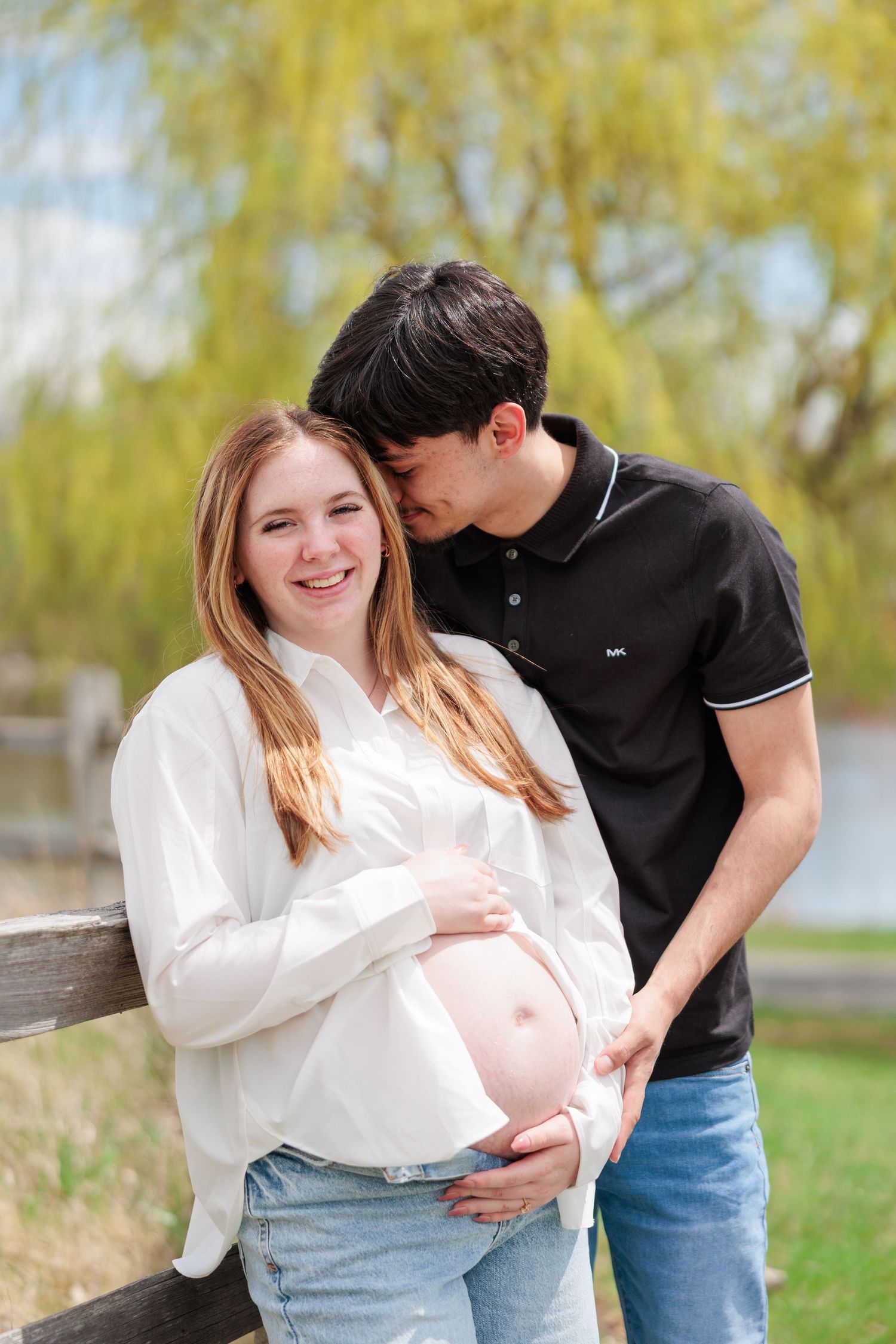 Smiling pregnant woman in white shirt and jeans with partner kissing her head near a wooden fence outdoors.