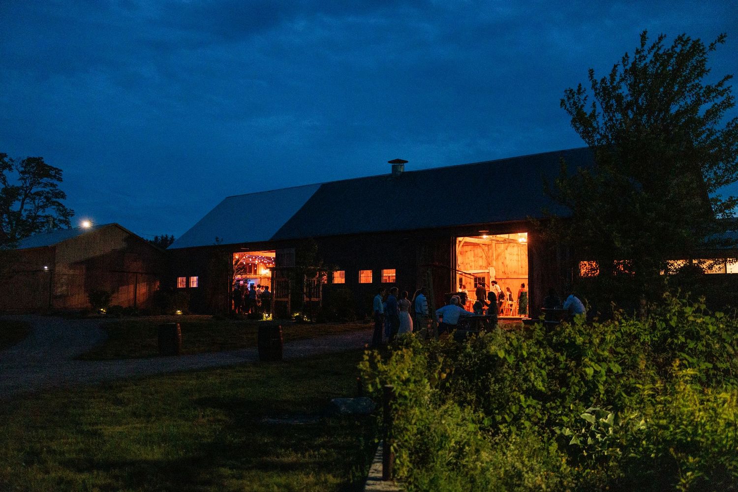 Rustic red barn wedding venue glows warmly at dusk with guests mingling outside under a deep blue sky.