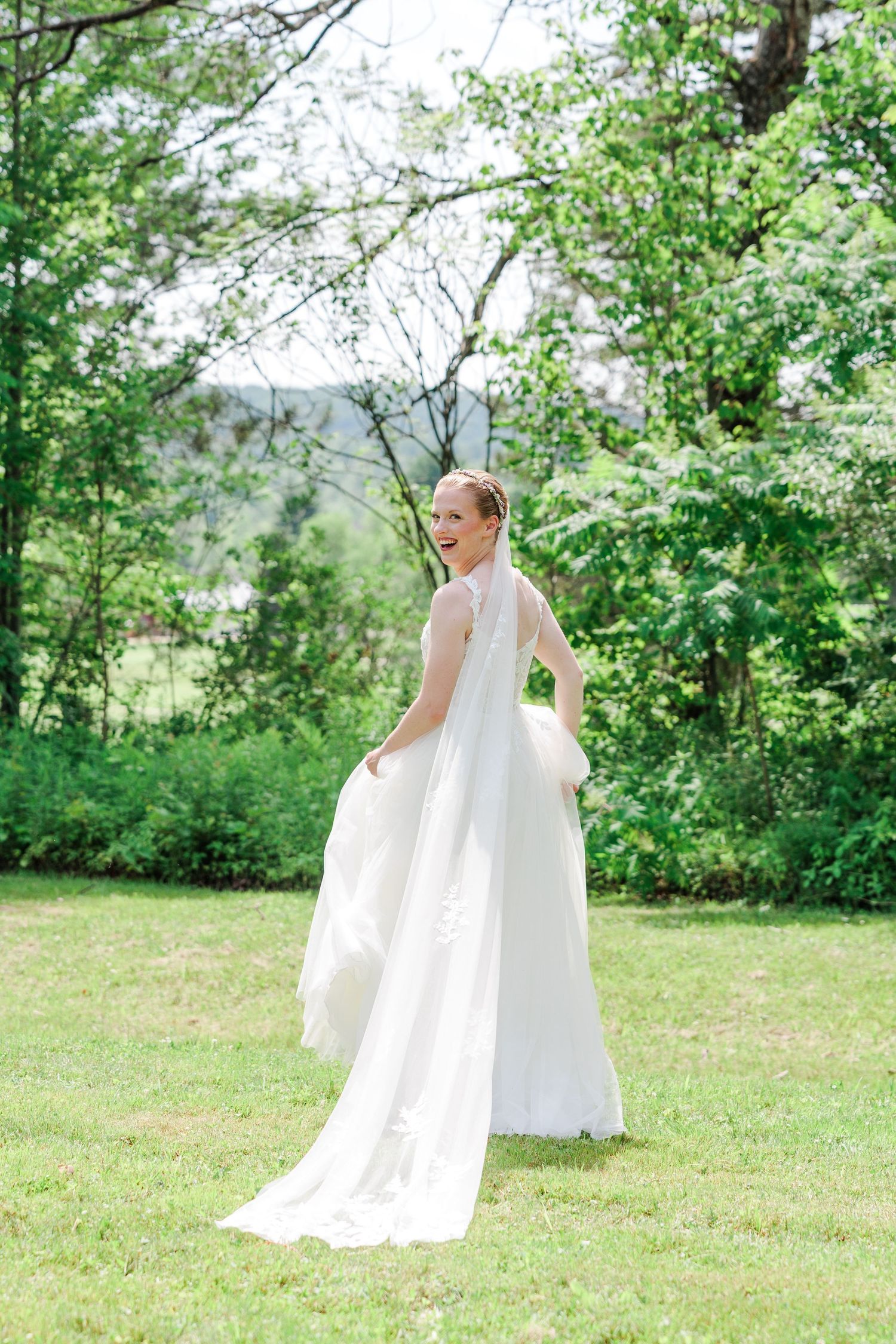 Bride in flowing white wedding gown twirling on green lawn surrounded by lush trees.