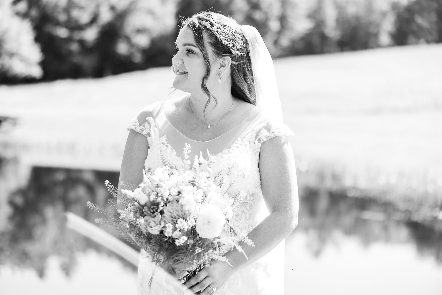 Bride in lace wedding gown holds bouquet outdoors in black and white portrait.