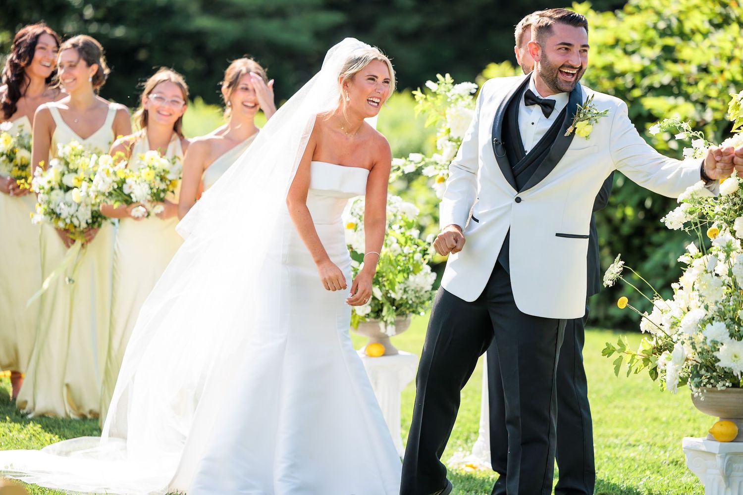 Jubilant groom in white tuxedo celebrates with bride in strapless gown after outdoor wedding ceremony.