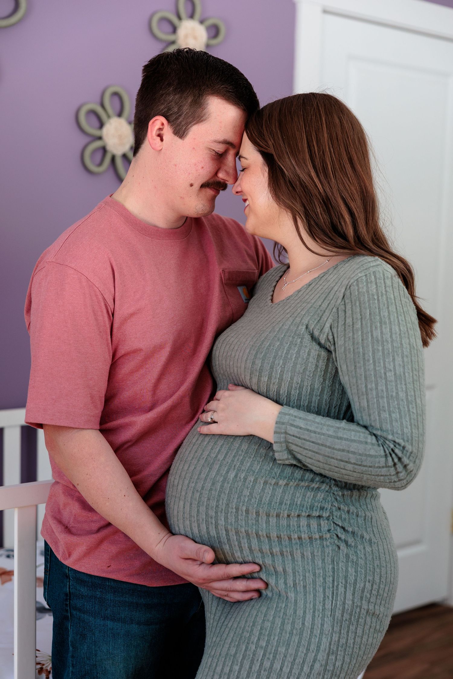 A loving couple touching foreheads while both cradling the pregnant woman's baby bump in a nursery.