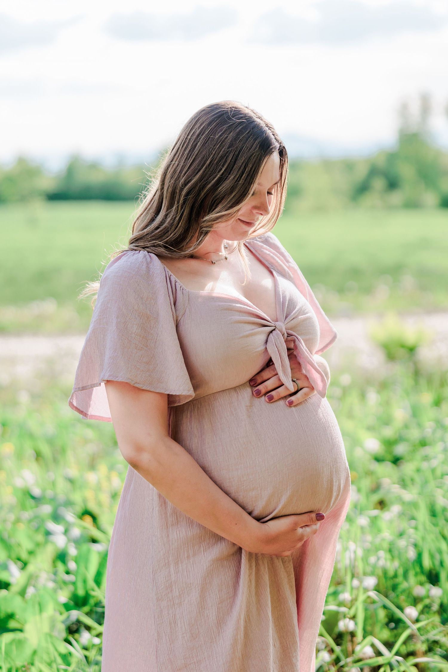 Pregnant woman in pink dress cradling her baby bump in a green field.