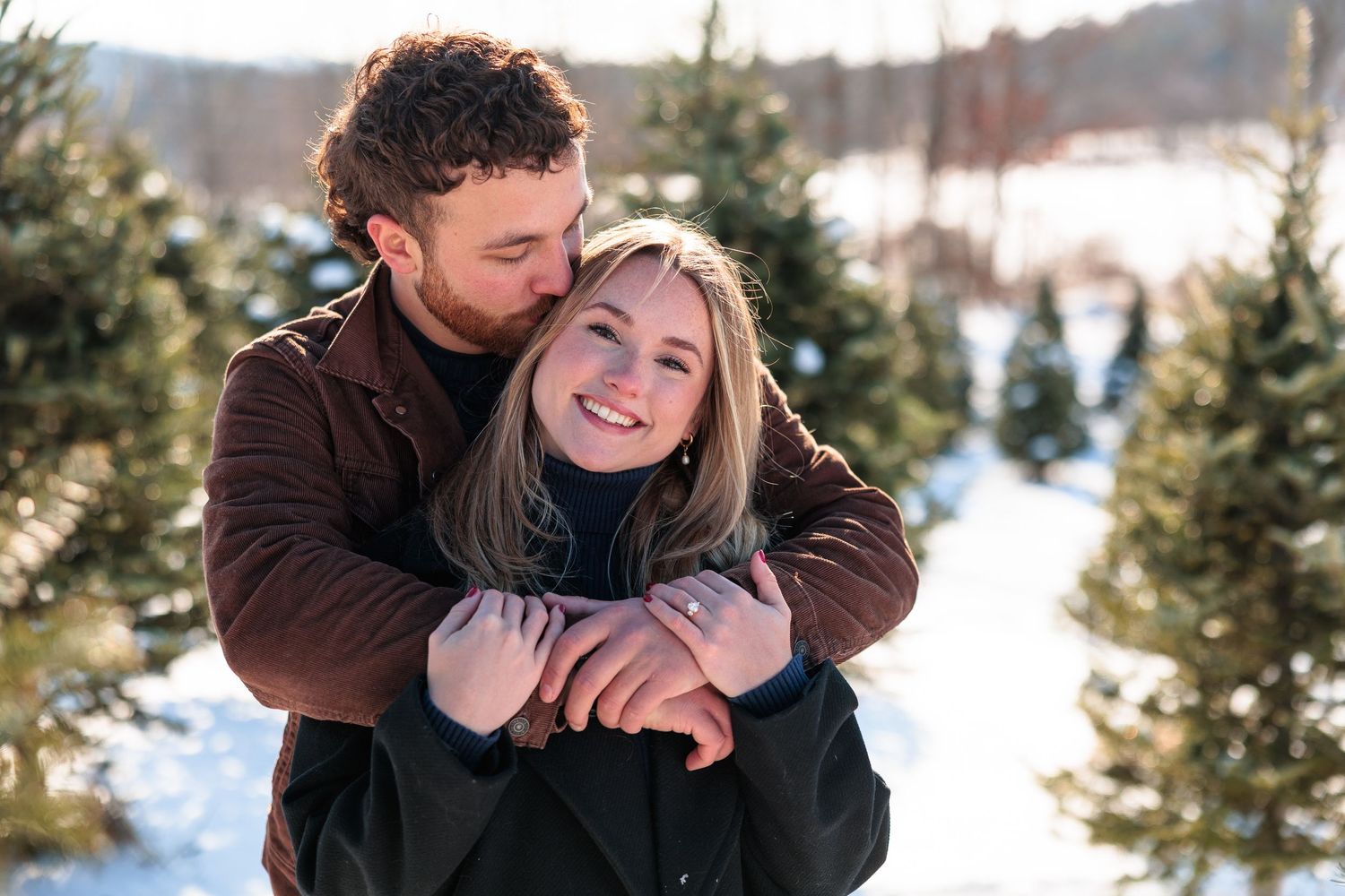 Man kissing woman on cheek while embracing her at a snowy Christmas tree farm.