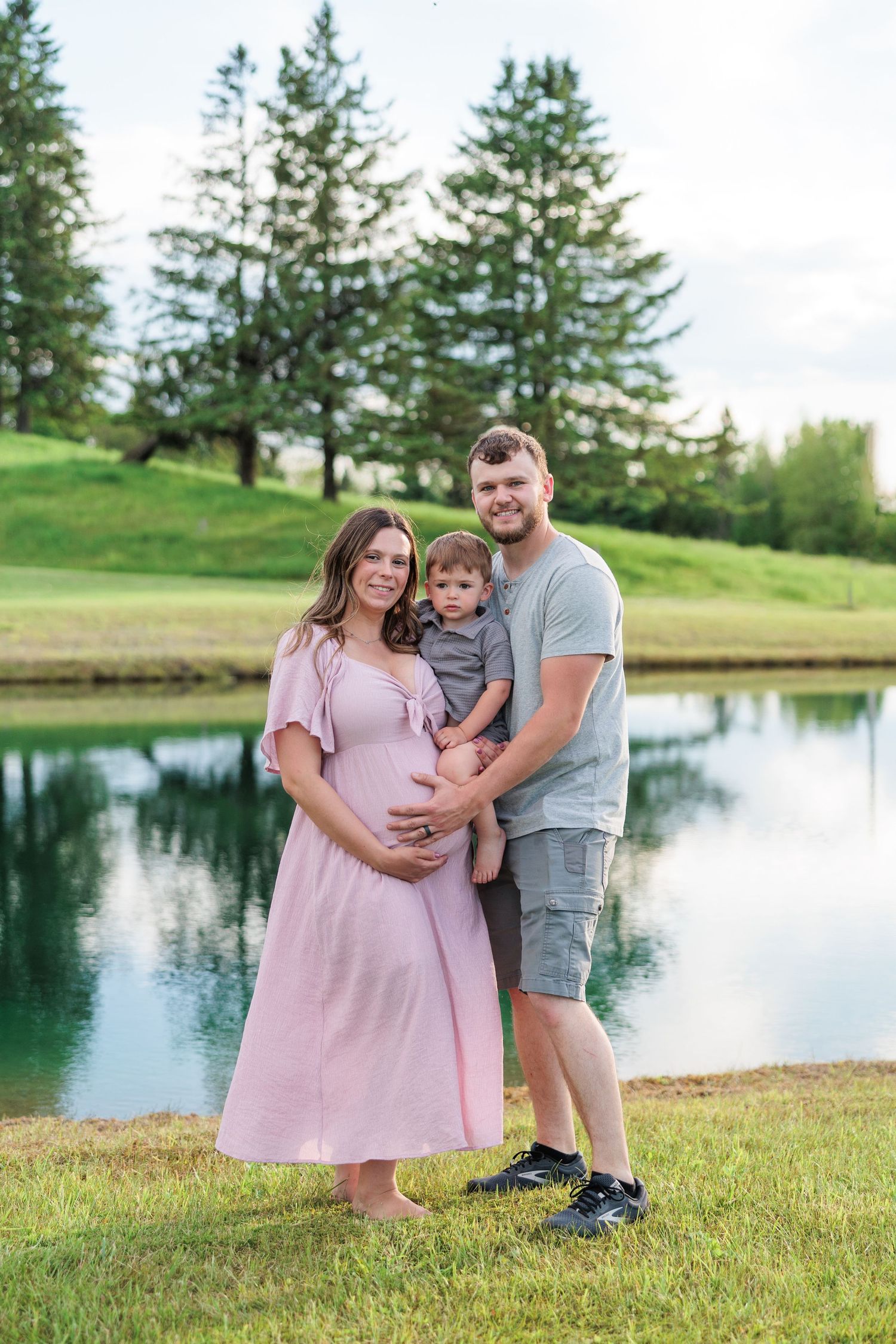 Happy family maternity photo with pregnant woman in pink dress, partner, and toddler by a serene pond.
