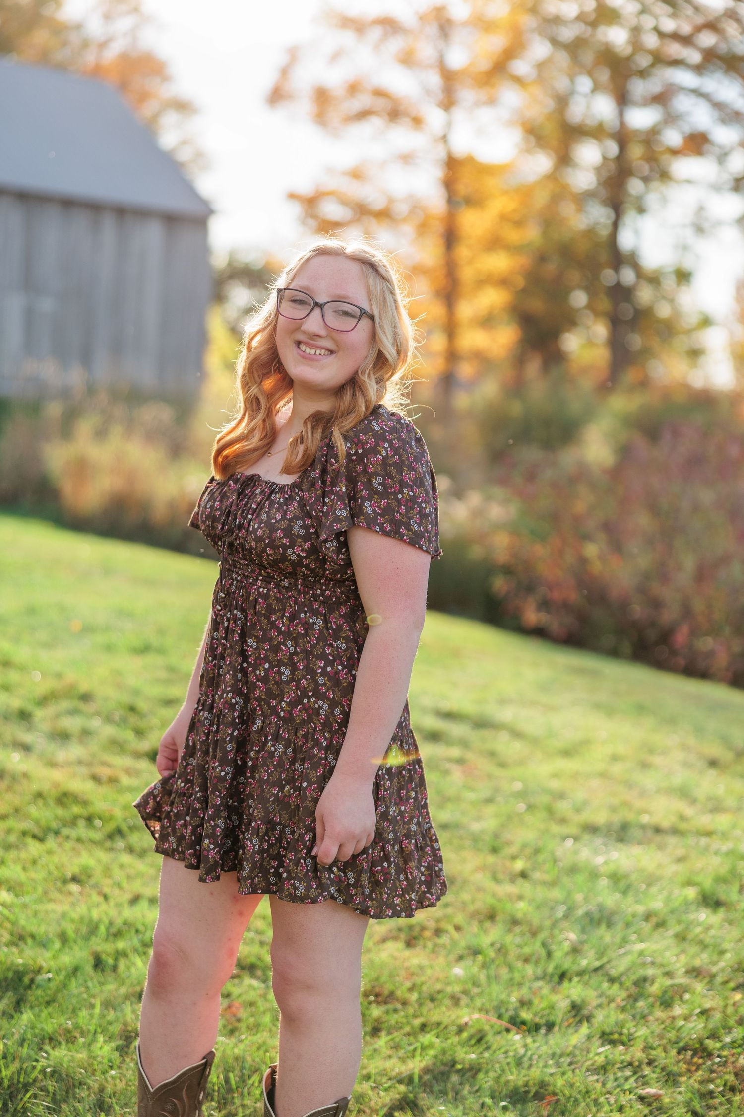 Young woman in floral dress and boots smiling outdoors in autumn setting with golden foliage and barn.