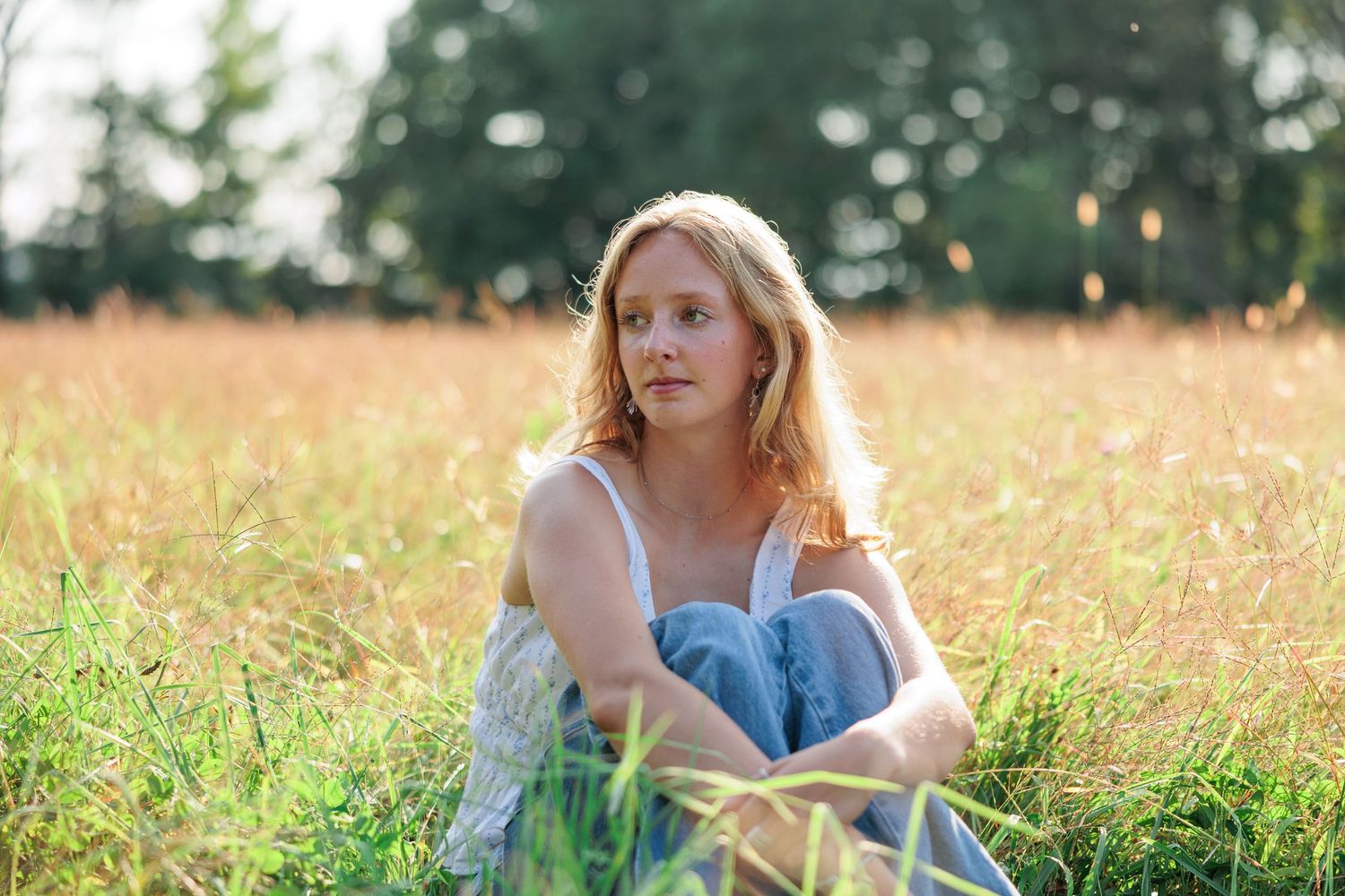 Young blonde woman sitting in sunny meadow grass wearing jeans and white top on summer day.