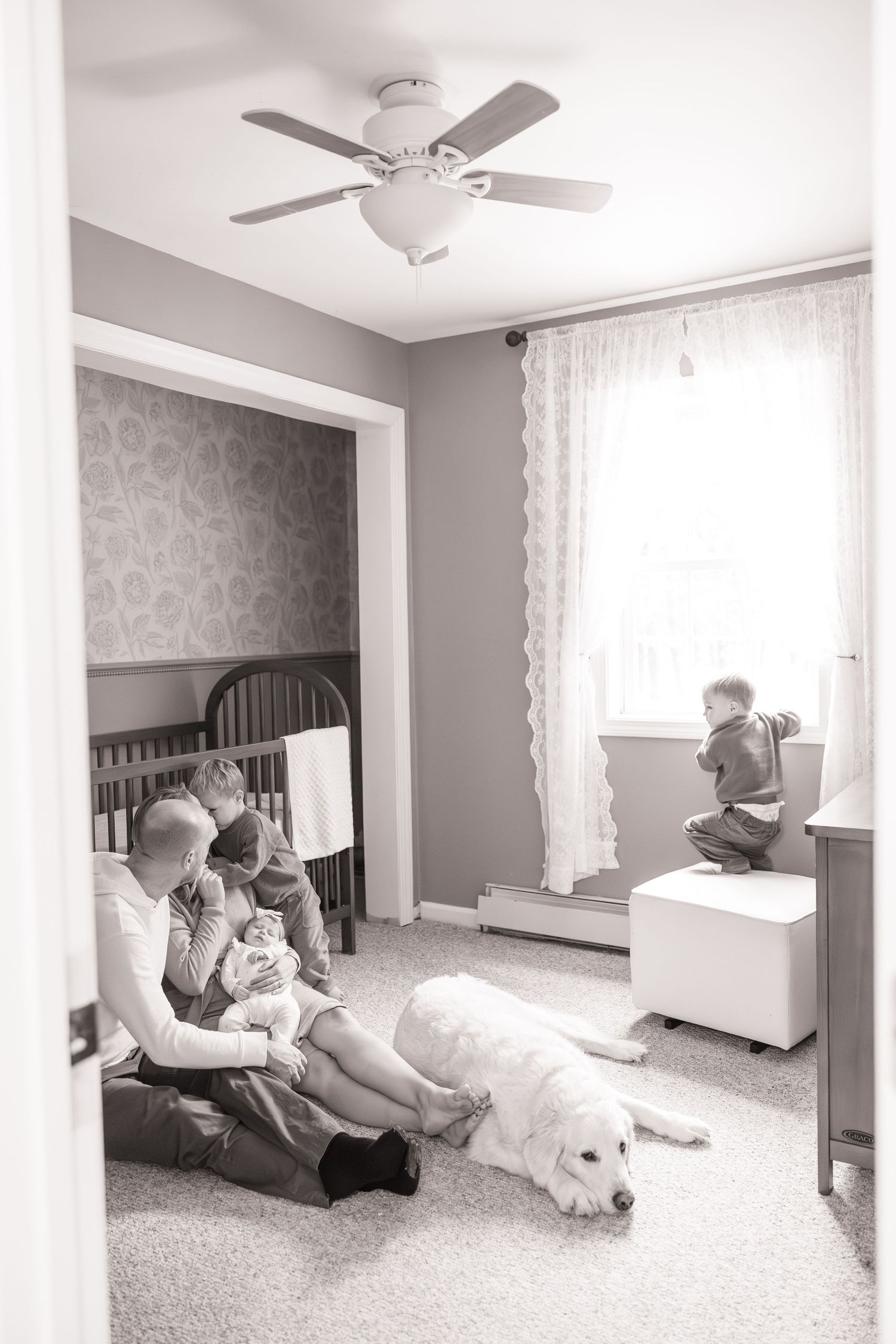 Black and white panoramic series showing intimate family moments in a nursery with ceiling fan and white curtains.