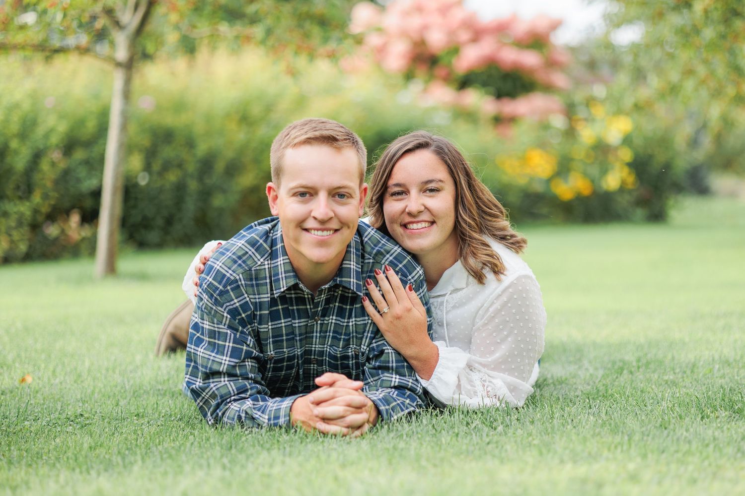Smiling engaged couple lying on green grass with woman showing off her engagement ring.